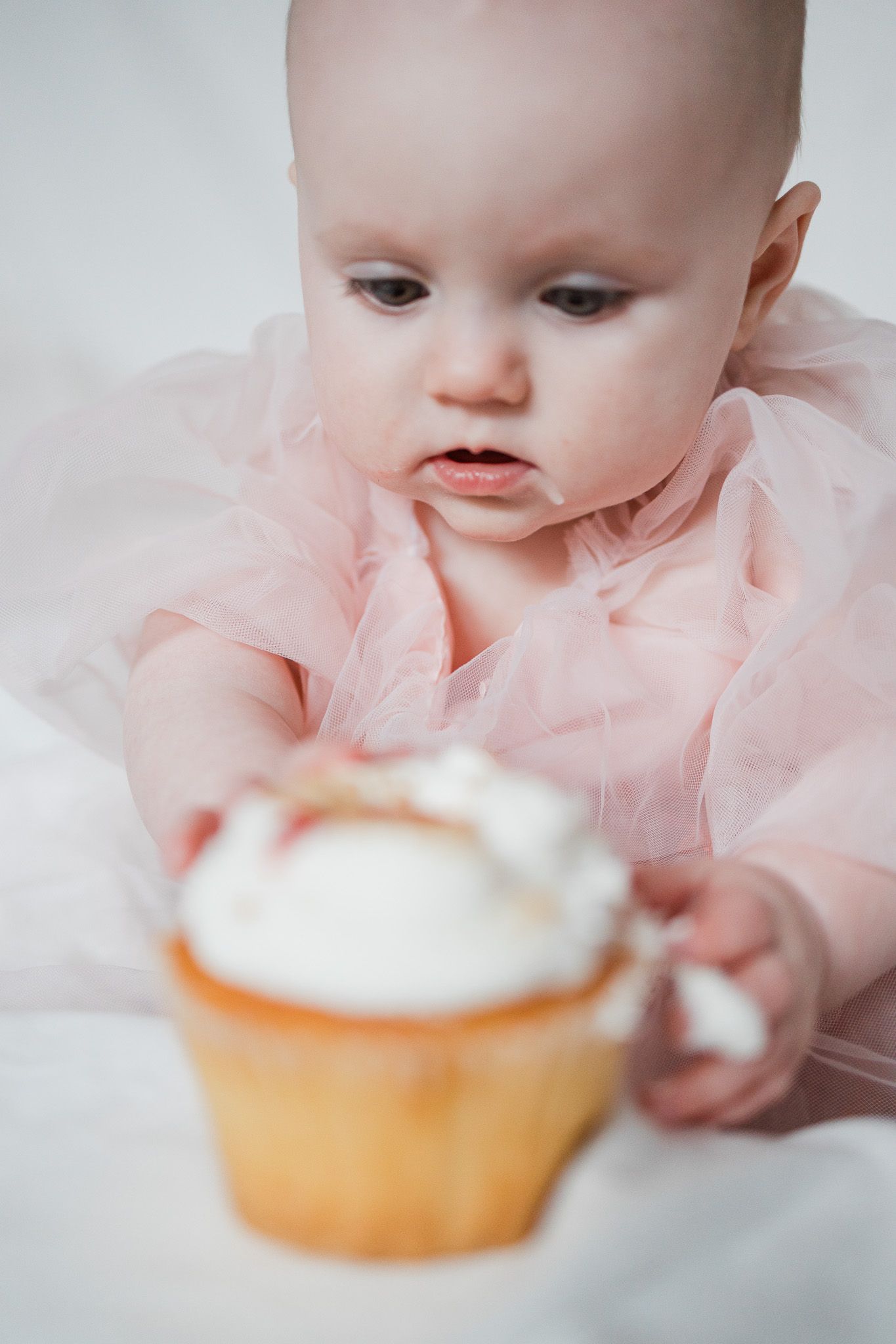 Baby in pink dress reaching for cupcake on white bedding.