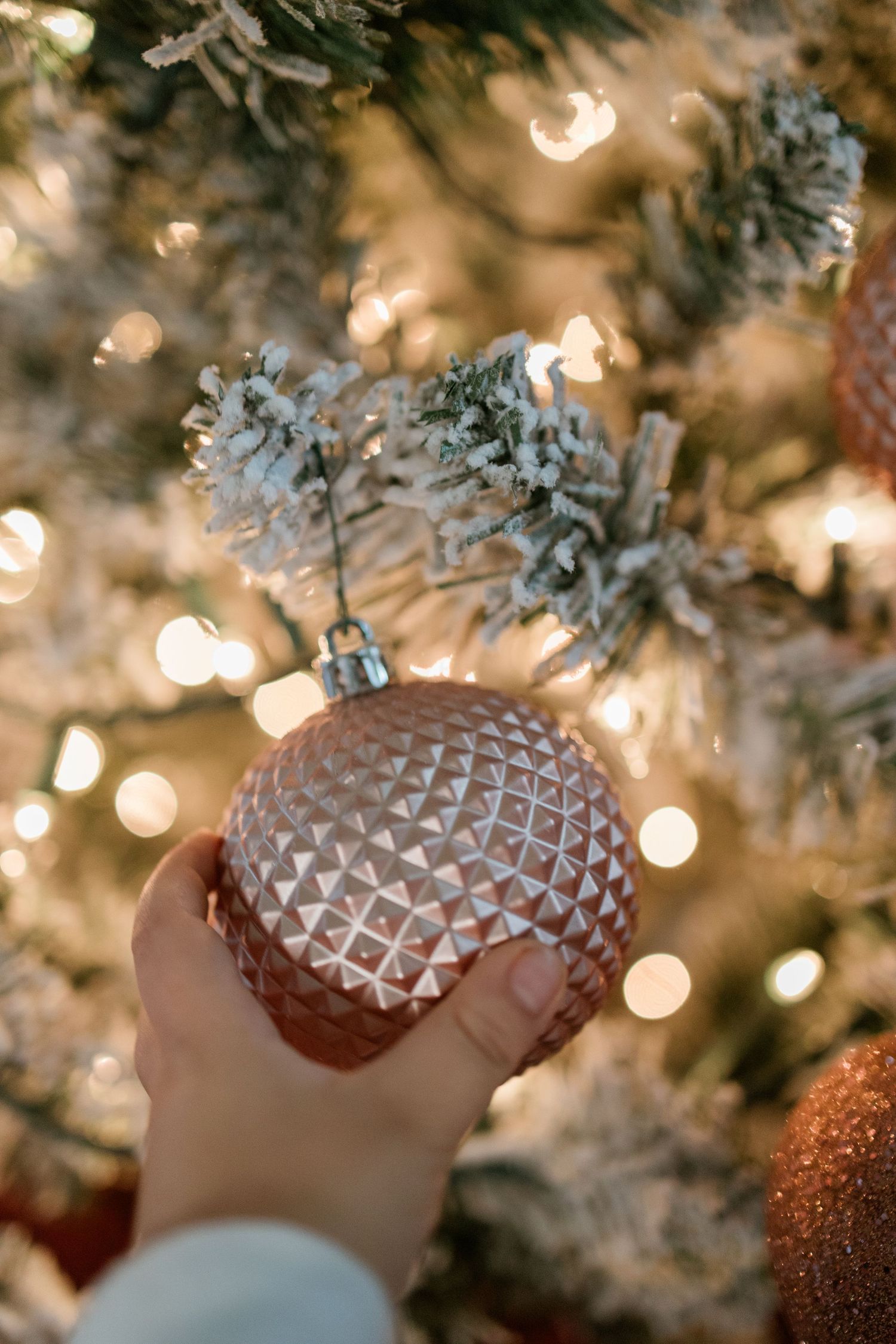 A hand holds a frosted glass Christmas ornament against a blurred background of twinkling holiday lights and snowy branches.