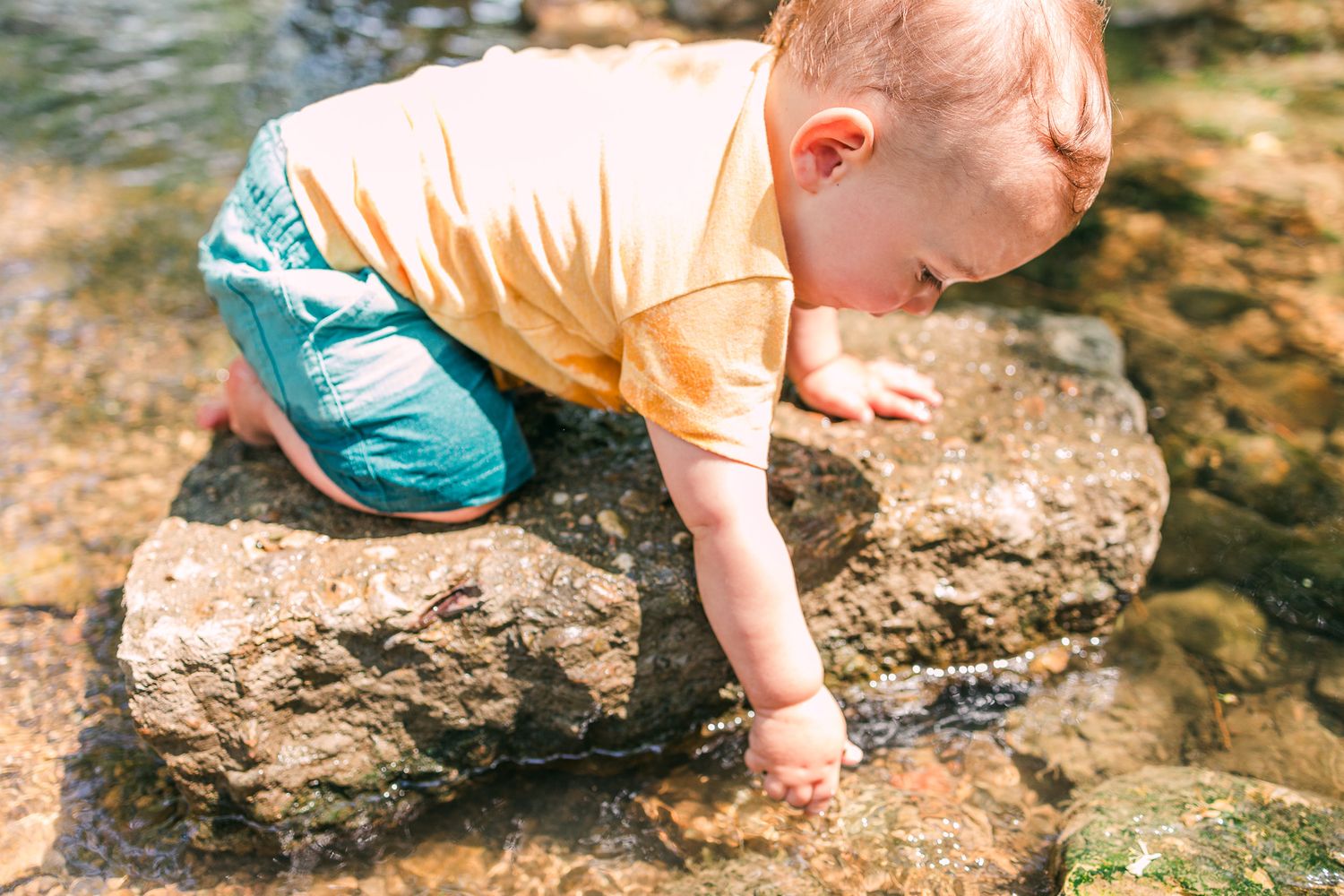 A small child in a yellow shirt and turquoise pants explores a rocky creek bed on a sunny day.