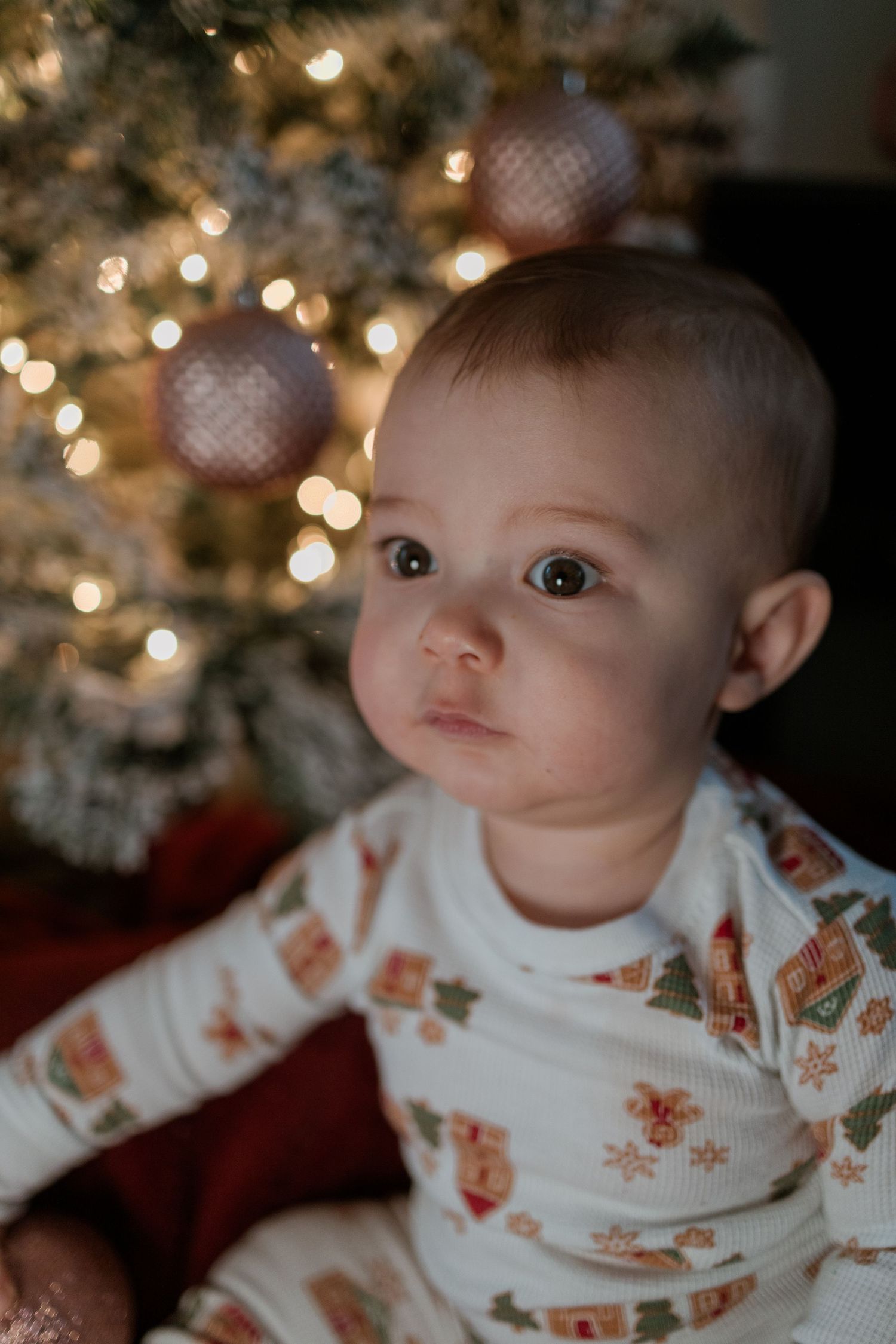 A baby wearing festive holiday pajamas sits near a lit Christmas tree with ornate ornaments and twinkling lights.