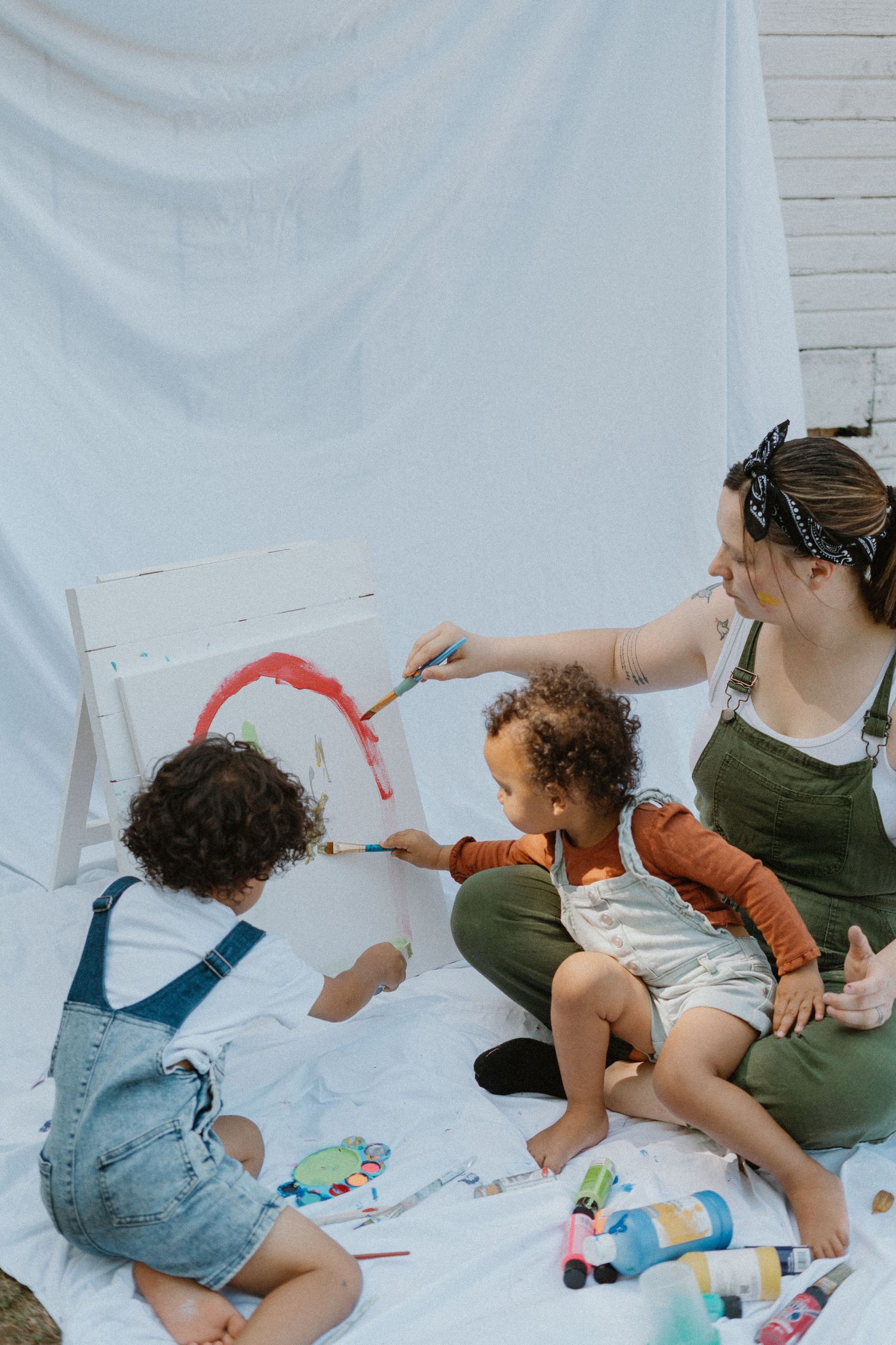 Children and caregiver painting together on white sheet with art supplies spread around.