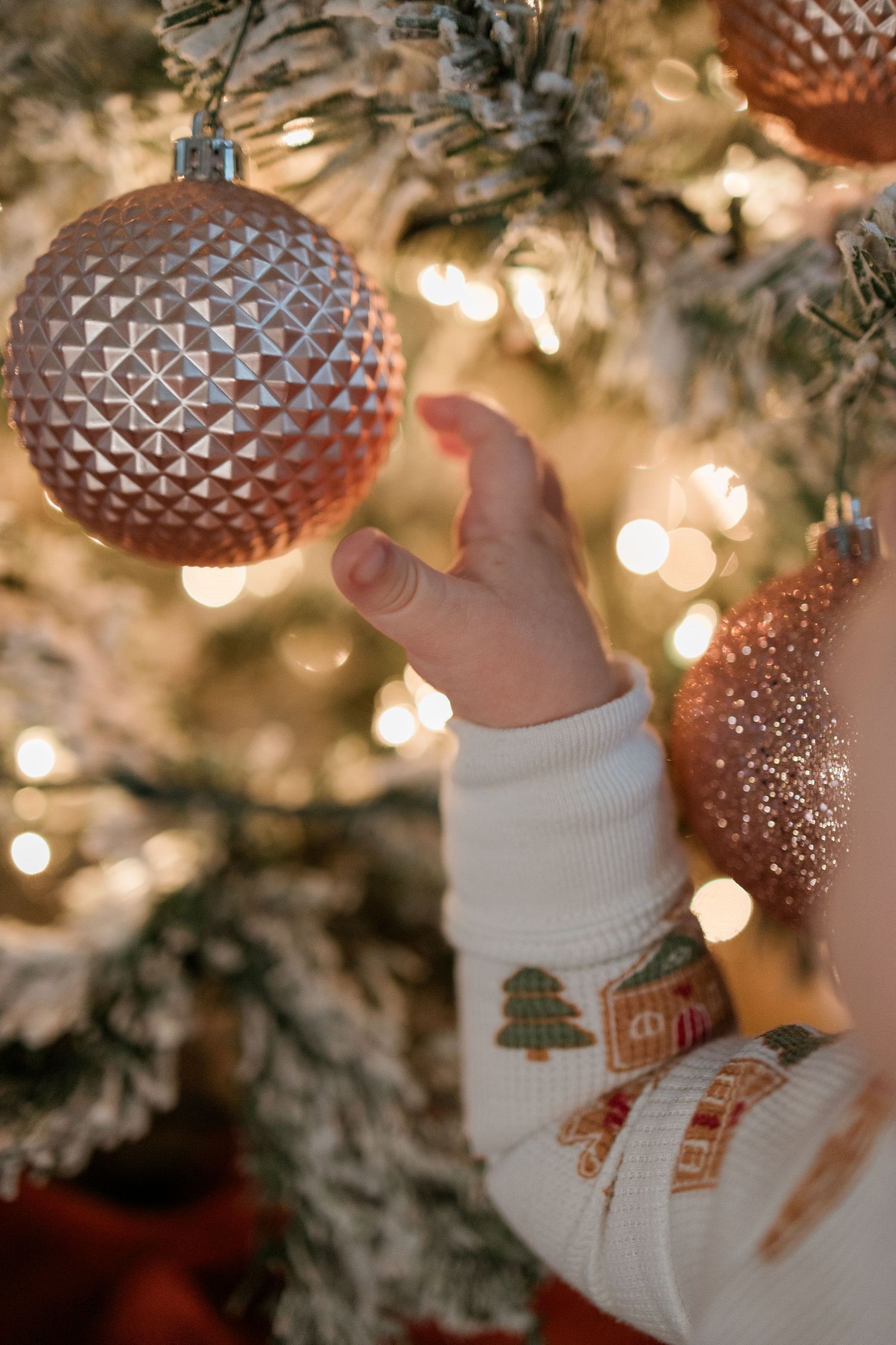 A hand in a holiday sweater reaches for a glittering rose gold ornament on a Christmas tree with twinkling lights.