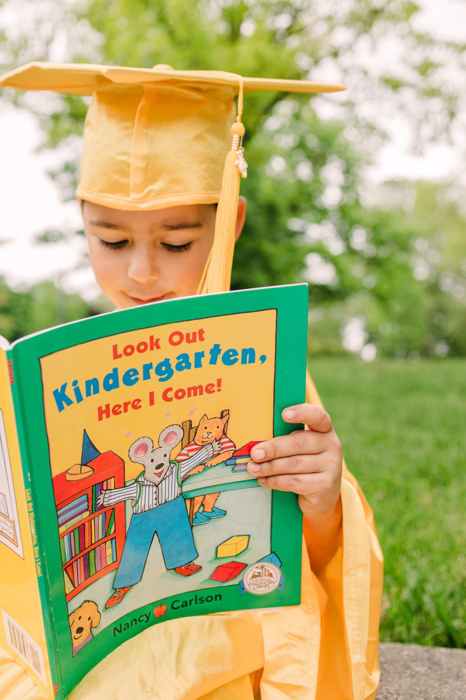 Graduate in yellow cap and gown reading 'Look Out Kindergarten, Here I Come!' book outdoors.