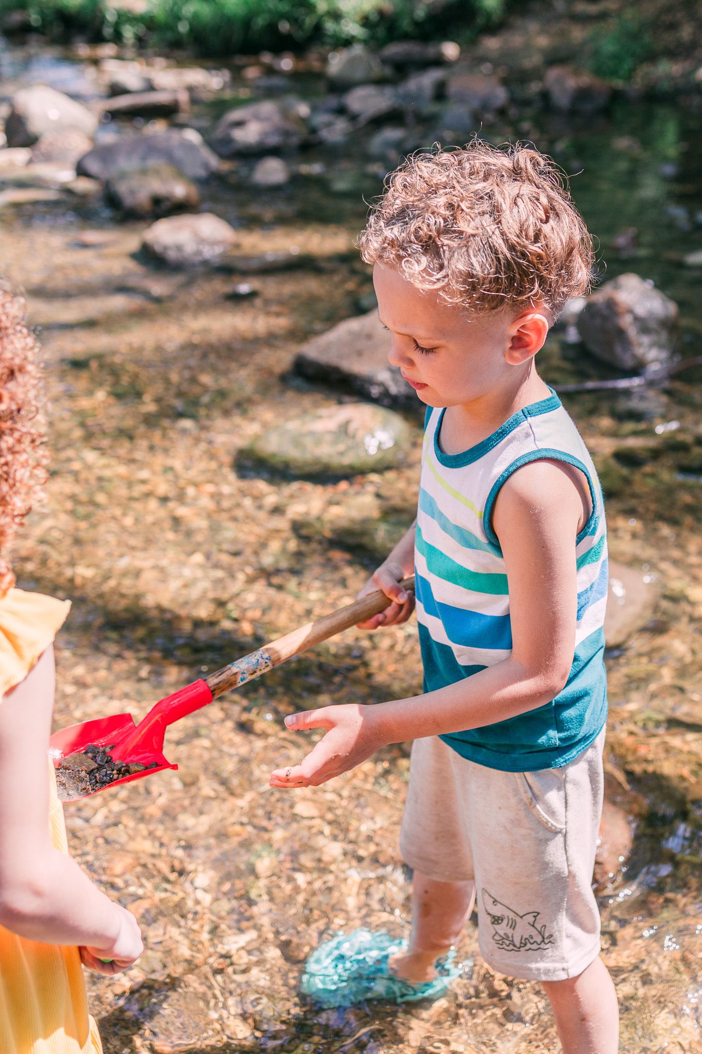 Young child in blue and white striped tank top plays outdoors on rocky terrain with sand and sticks.