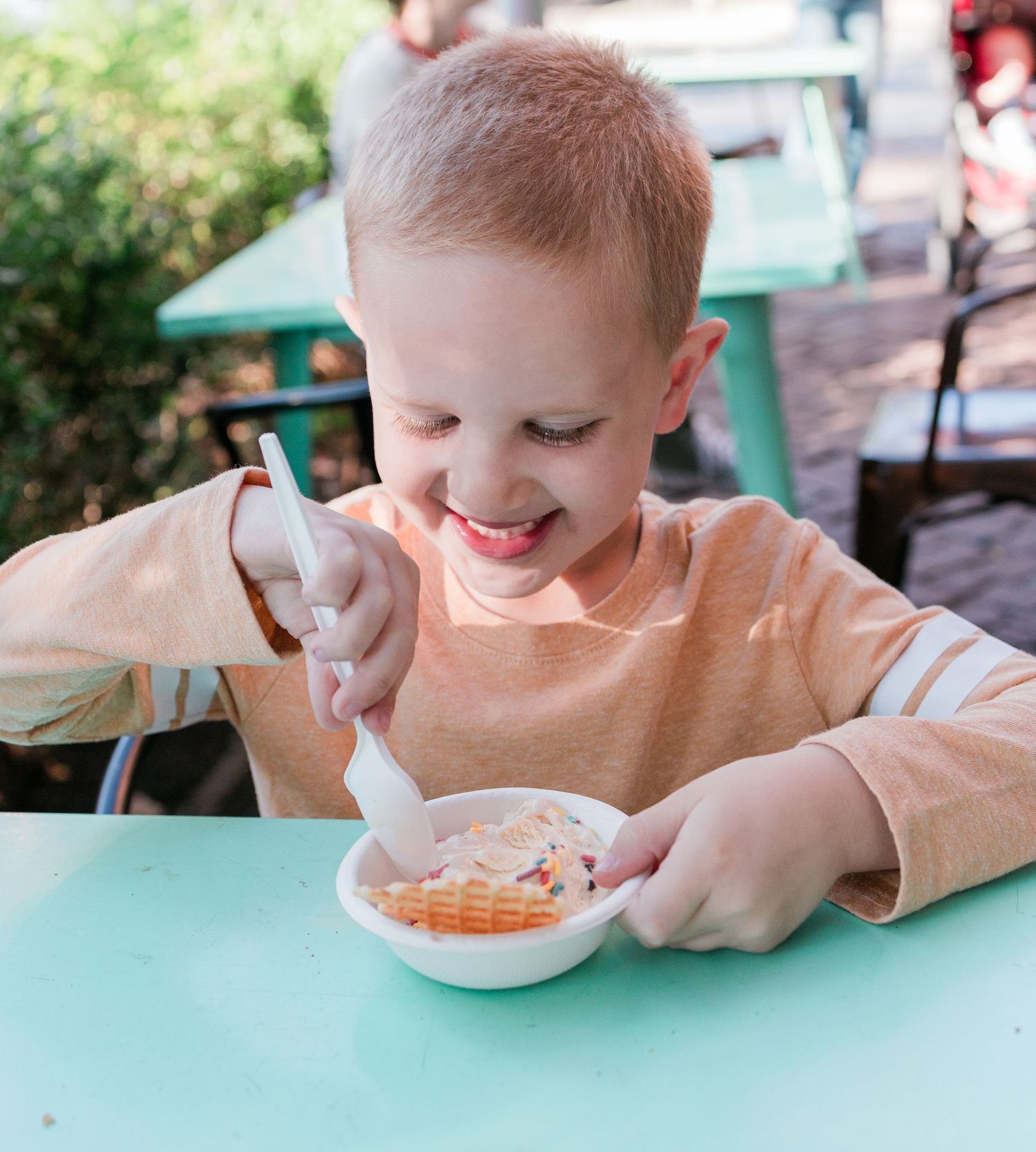 A child smiles while eating from a small bowl at a turquoise table outdoors.