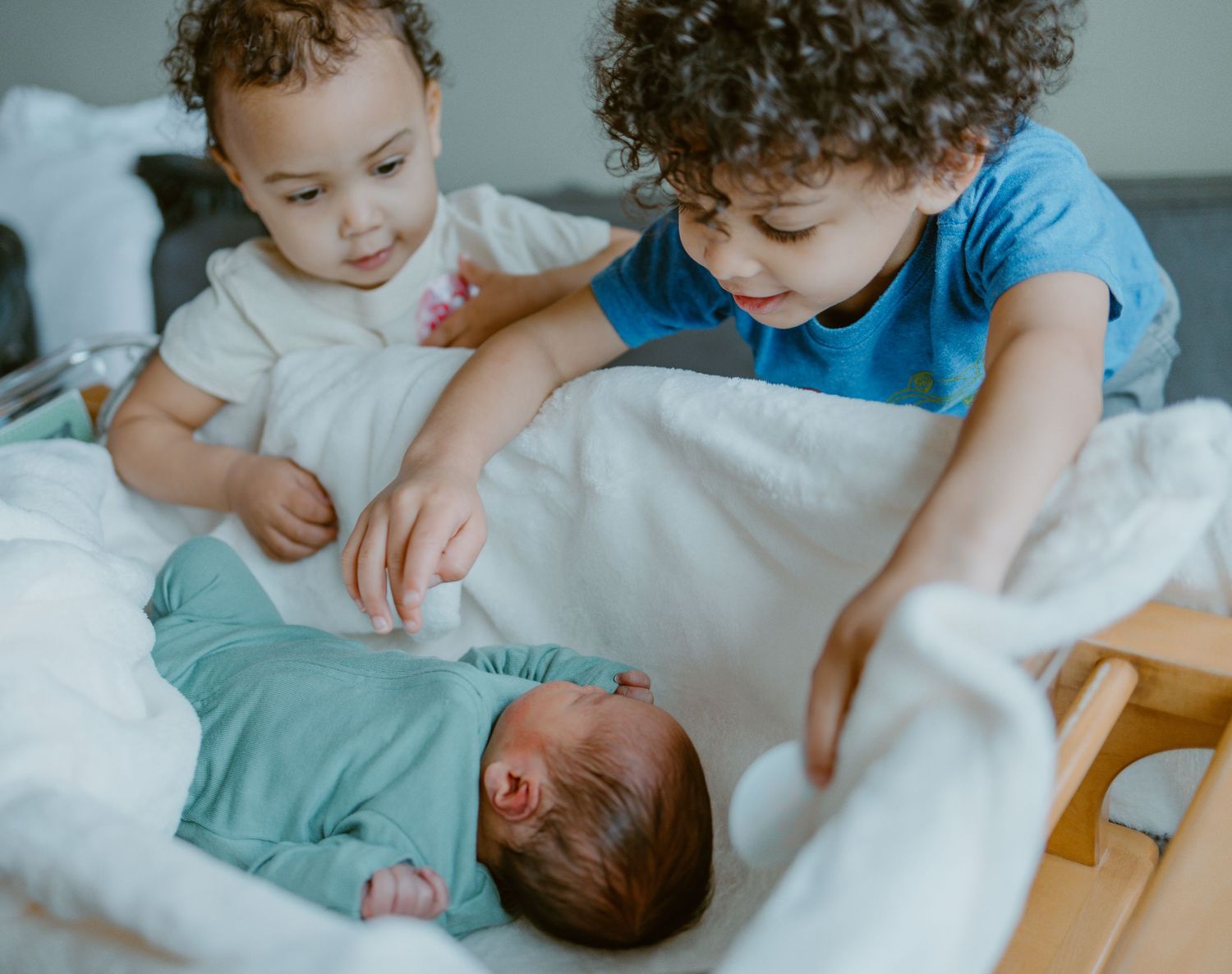 Young children gather around to gently greet and touch a newborn baby lying on white bedding.