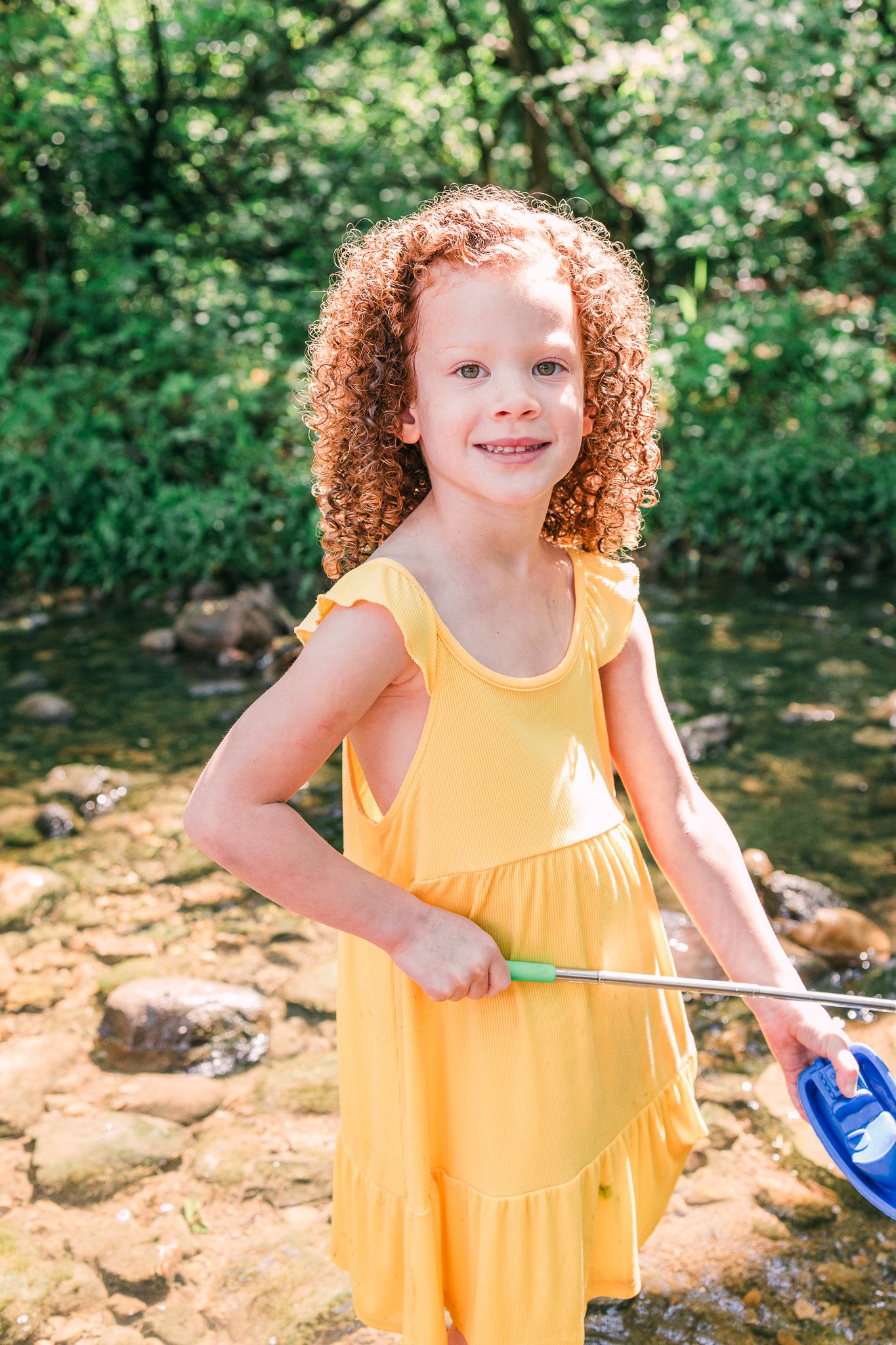 A child in a yellow dress stands creekside on a summer day, holding a blue item against a lush green background.