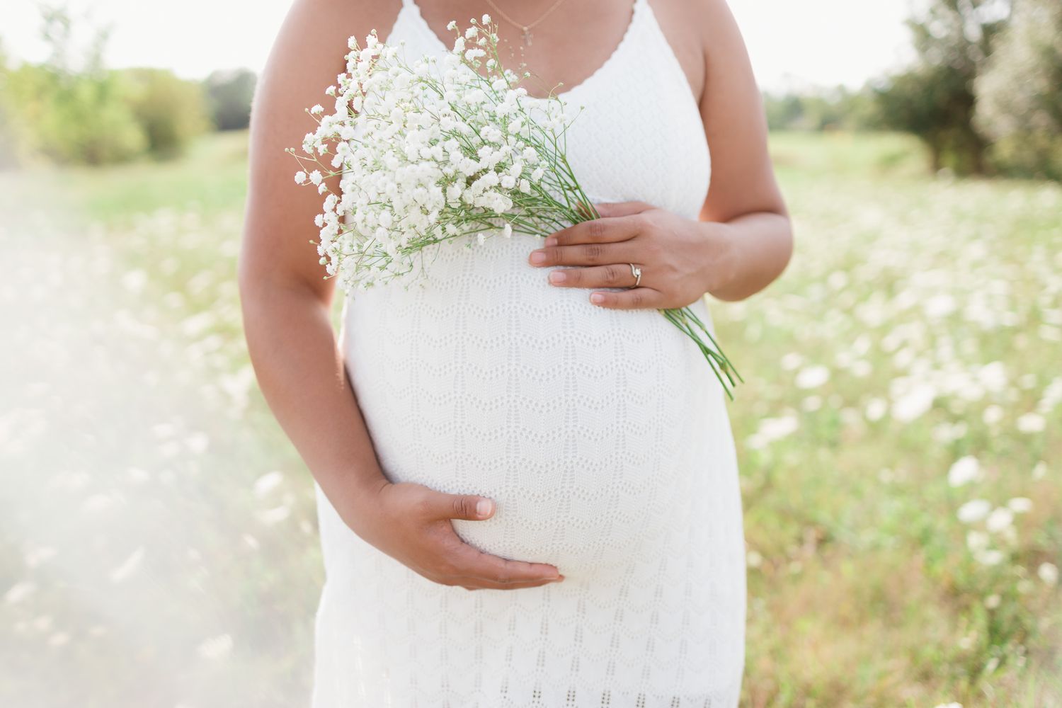A pregnant person in white dress holds delicate baby's breath flowers against their belly outdoors.
