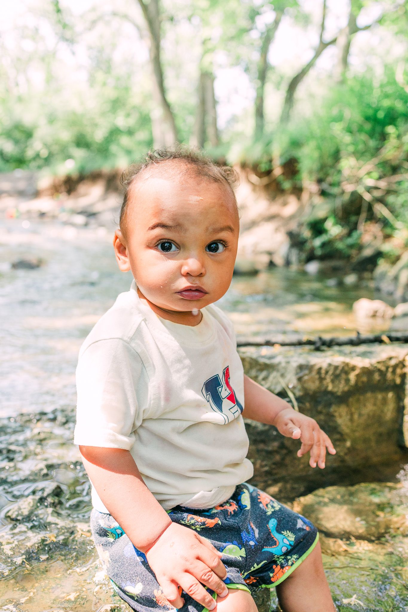 An infant in a white t-shirt and patterned shorts sits on a stone path surrounded by greenery in a natural outdoor setting.