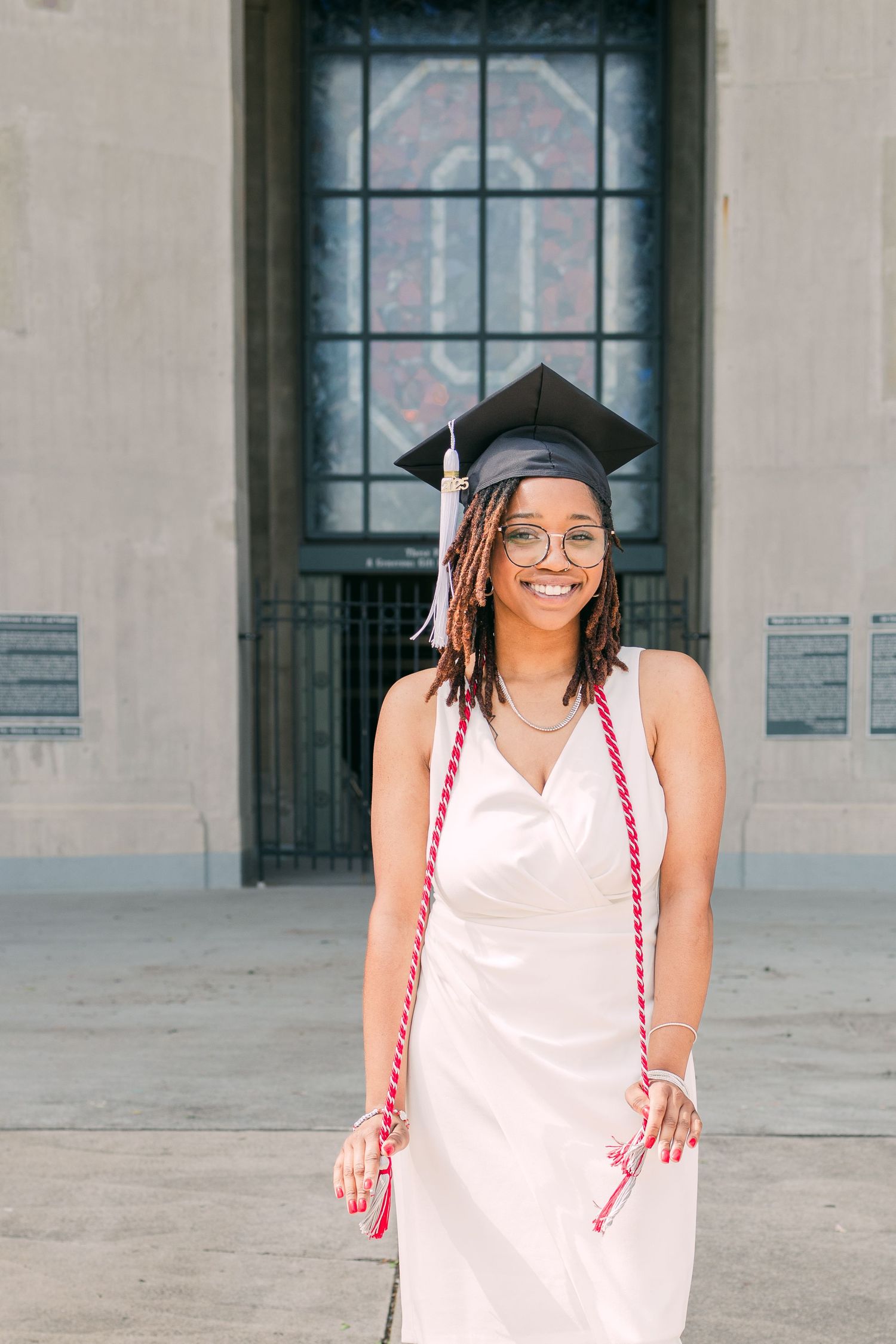 Graduate in white dress and cap posing in front of historic building entrance.