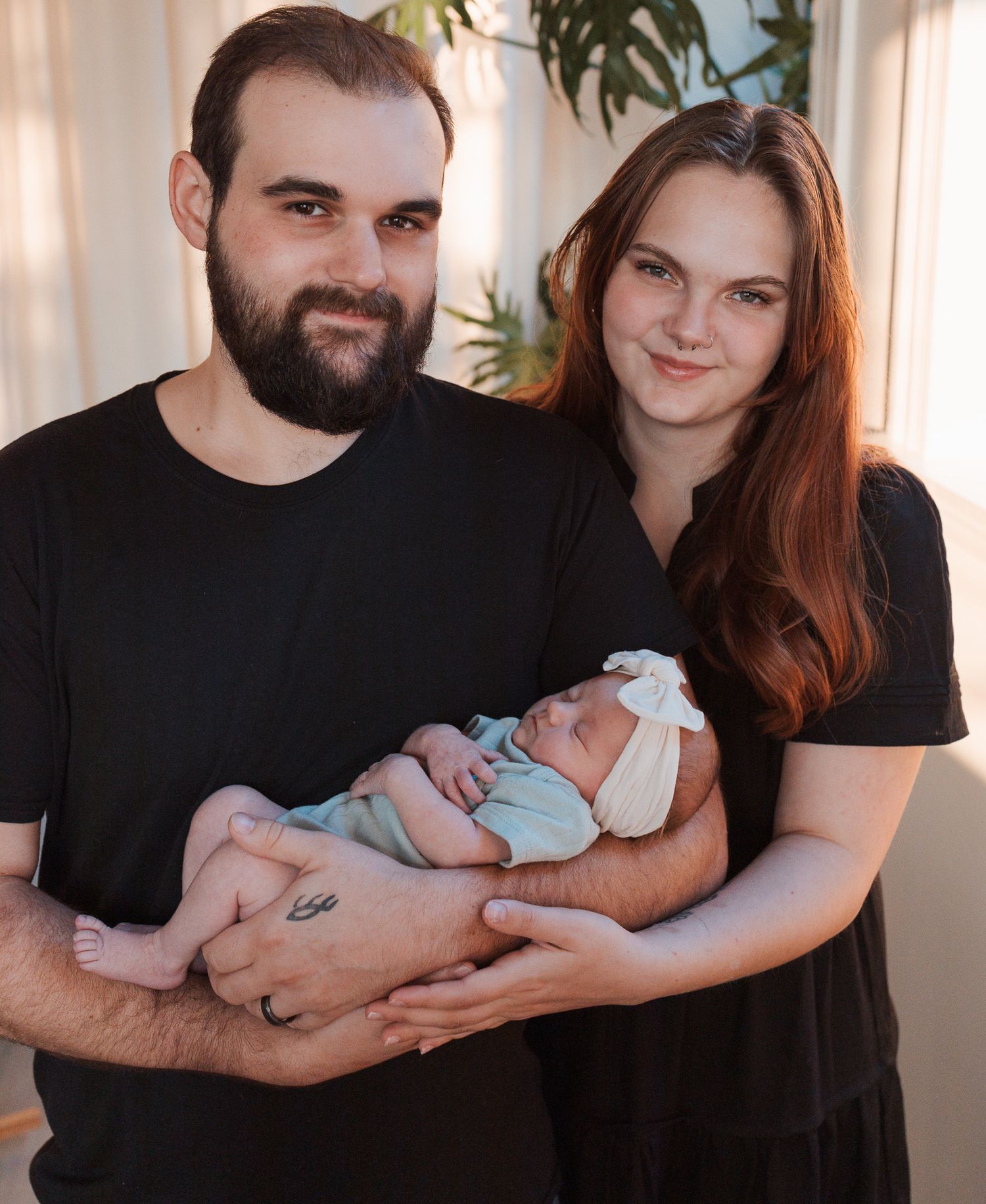 A couple in matching black outfits holds their newborn baby in a light blue onesie near a sunny window.