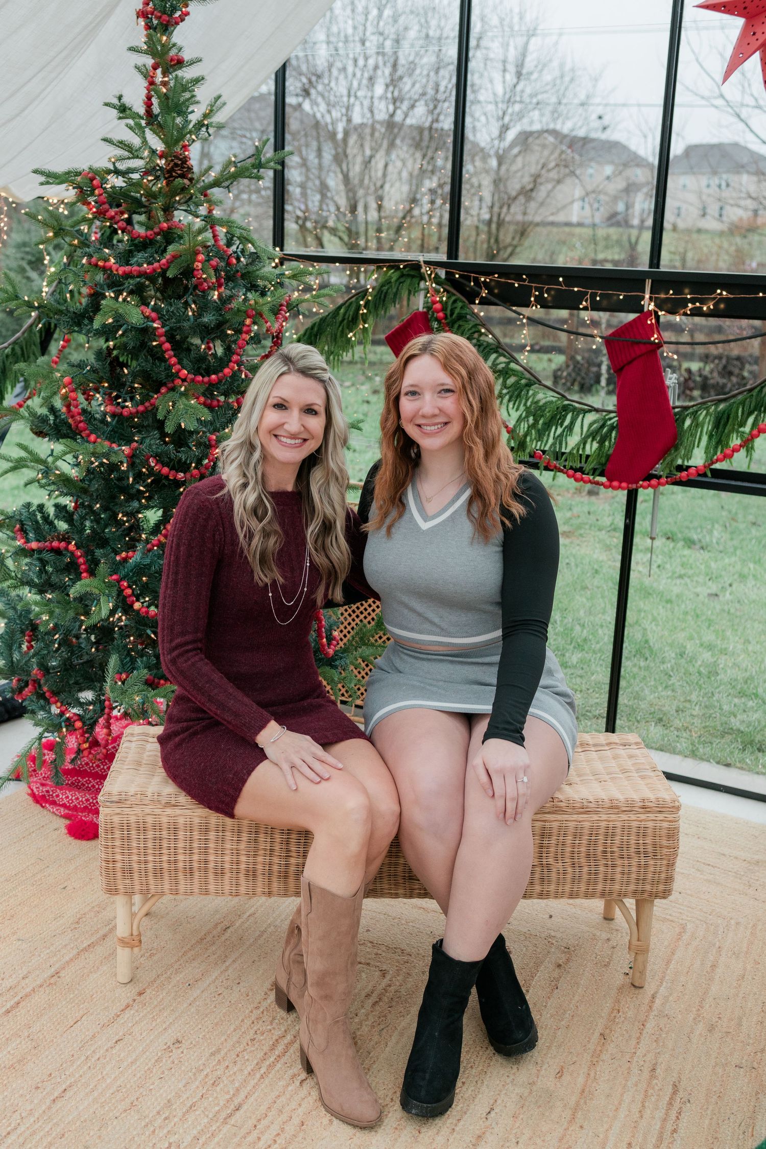 Two women in sweater dresses sit on a wicker bench by a decorated Christmas tree in a sunroom.
