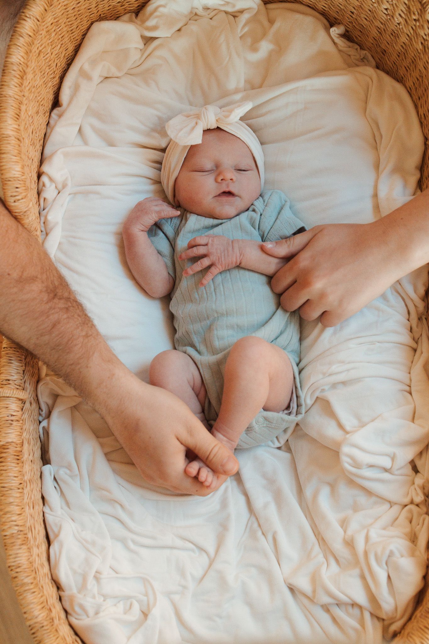 Sleeping infant wearing mint green onesie and bow headband rests peacefully in woven bassinet with white bedding.