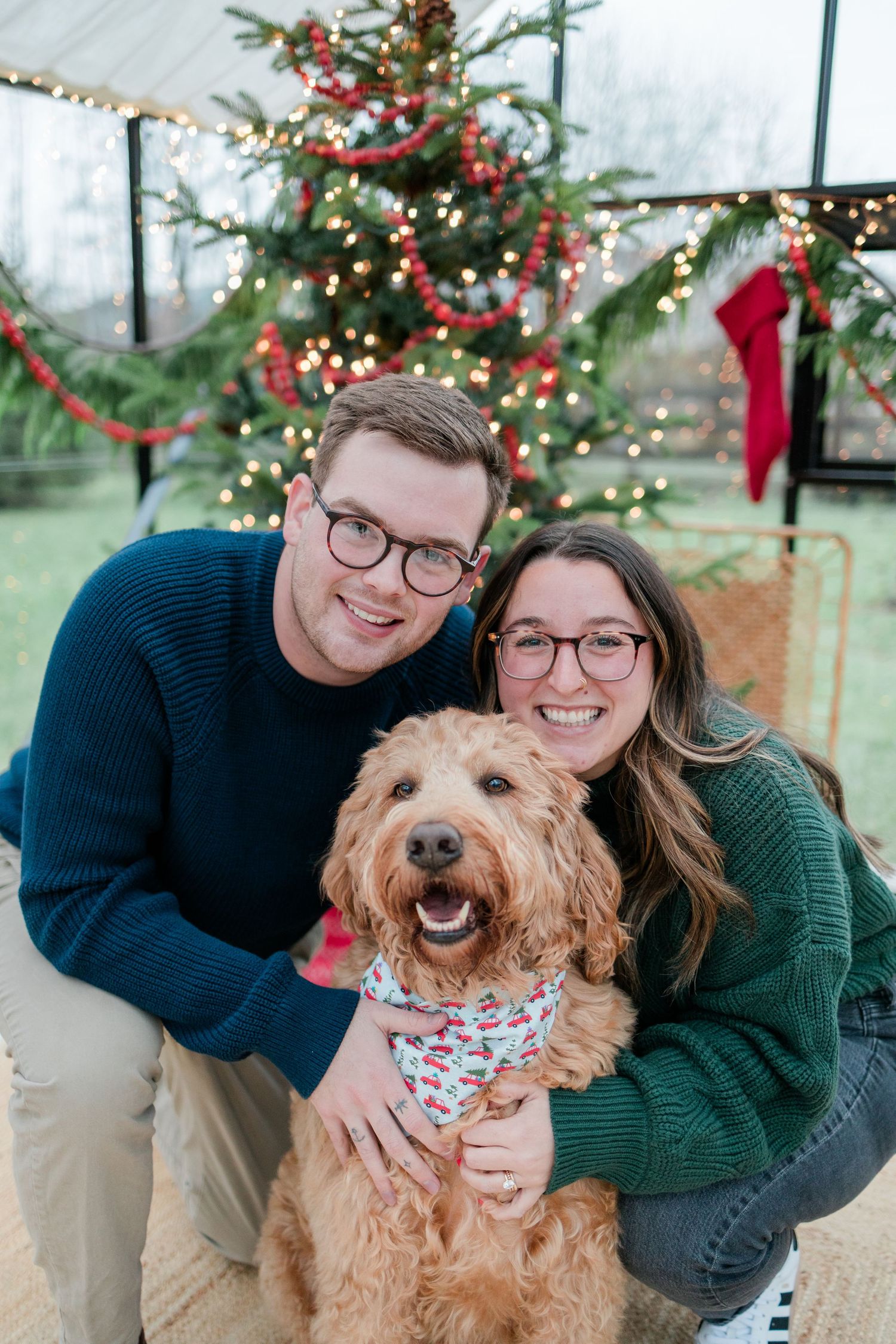 Couple and their goldendoodle dog pose together by decorated Christmas tree.