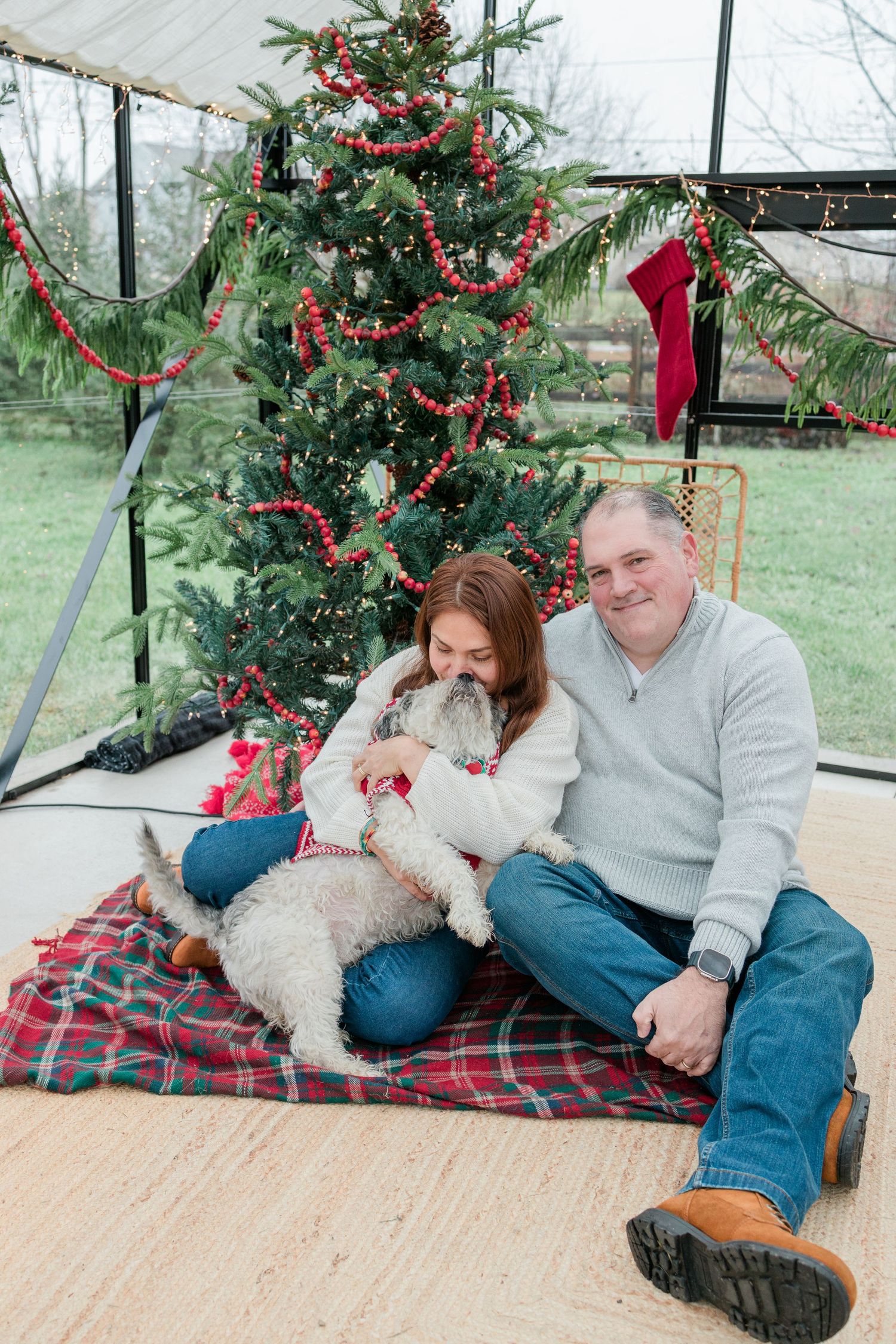 Couple and dog sitting on plaid blanket by Christmas tree in sunroom with festive decorations.