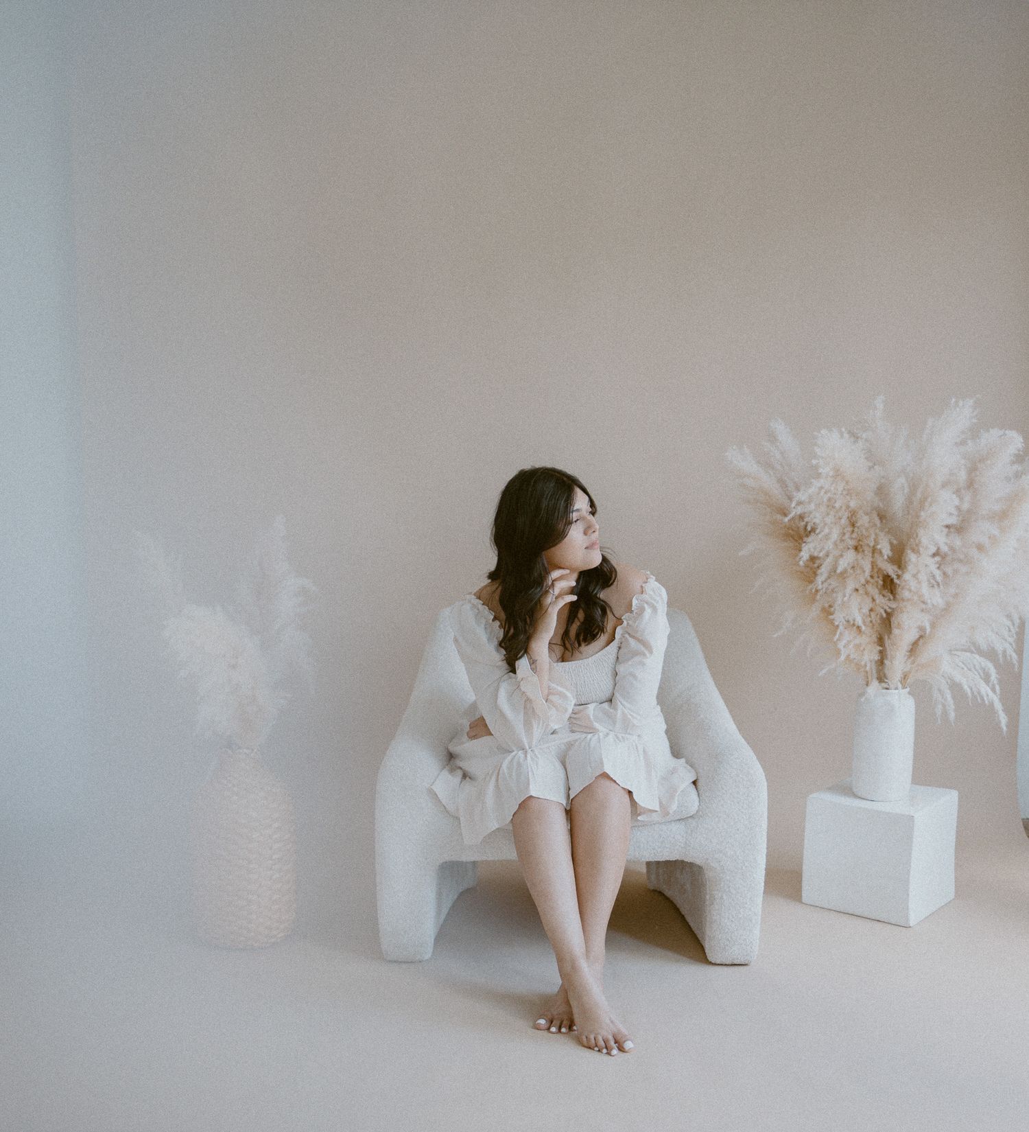 A minimalist portrait showing someone in white lounging beside pampas grass arrangements on white.