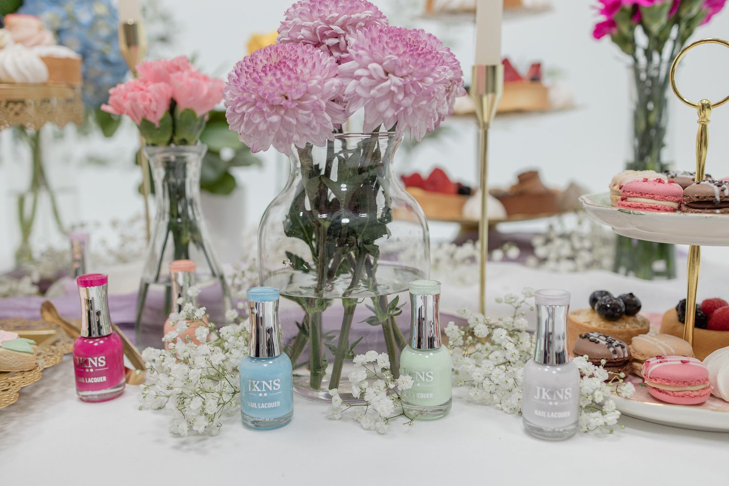 Pink floral centerpiece with nail polish bottles and tiered dessert displays on white table.