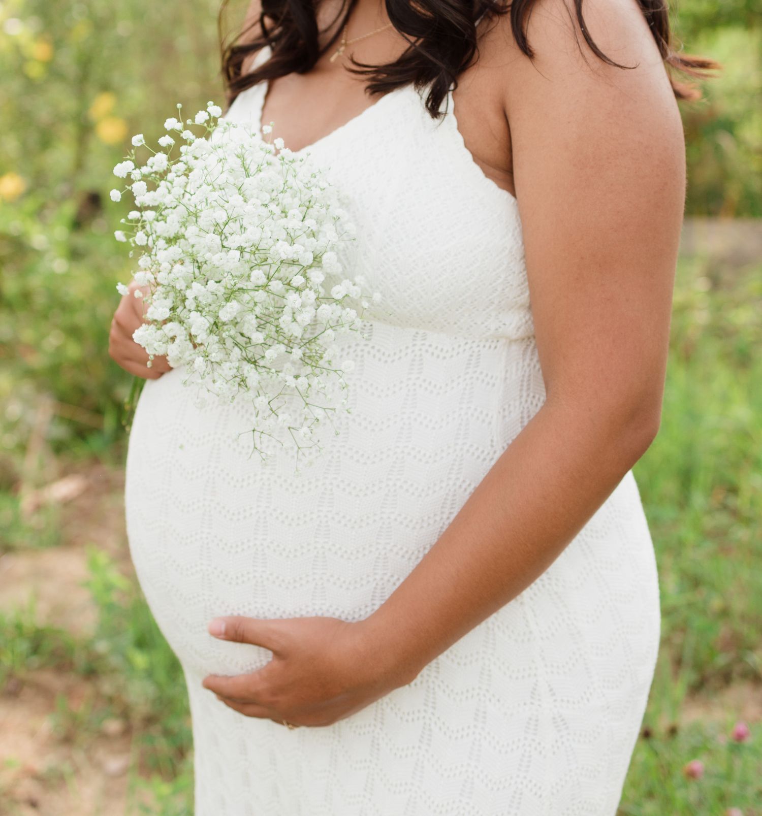Expectant mother in white dress holds delicate baby's breath flowers against her belly.