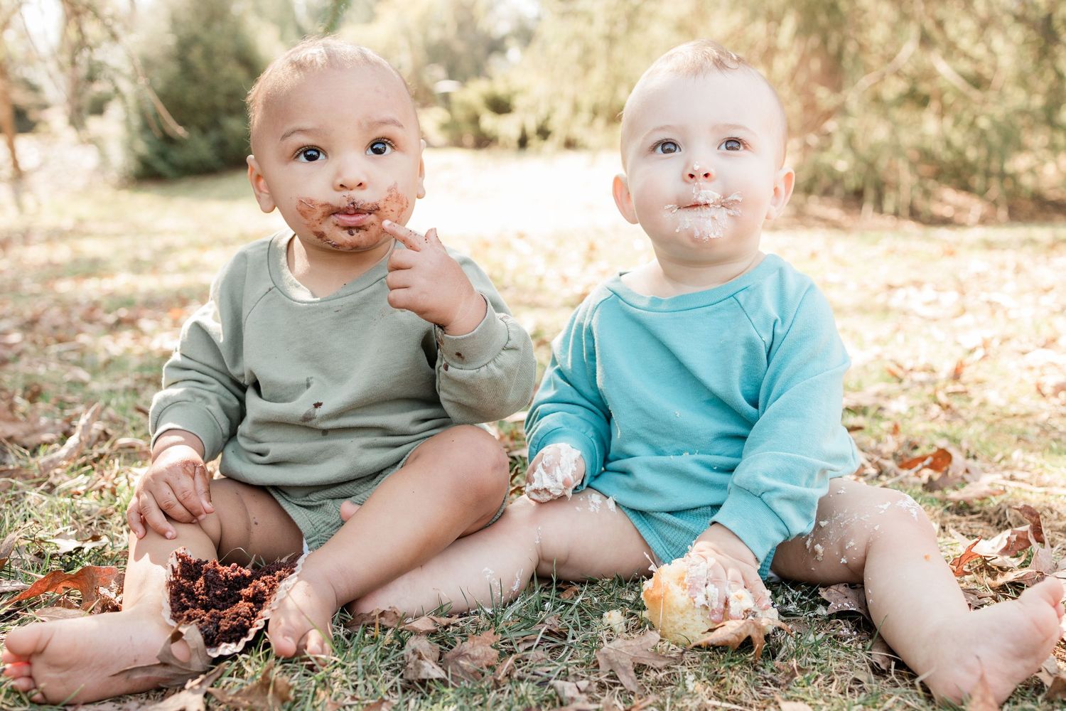 Two babies in matching outfits sitting on grass with messy faces from eating cake.