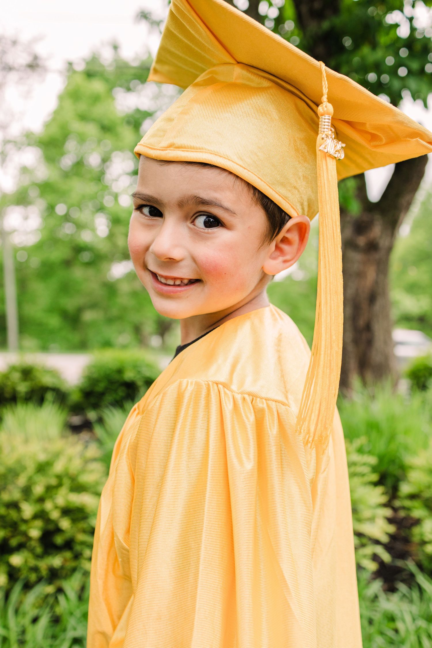 Graduate in yellow cap and gown smiling over shoulder in garden setting.