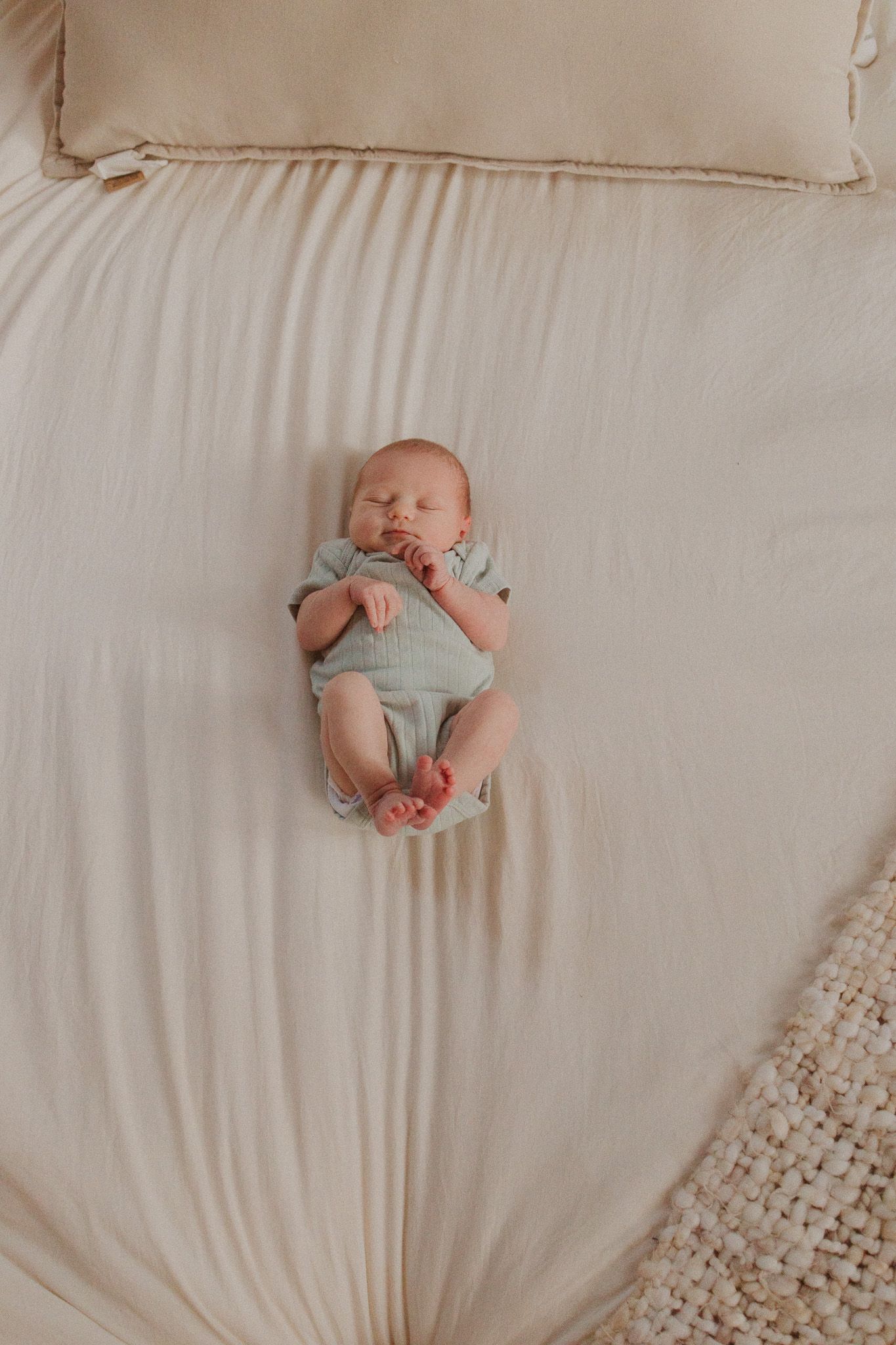 A newborn baby dressed in light clothing lays on white bedding in a series of minimalist lifestyle photos.