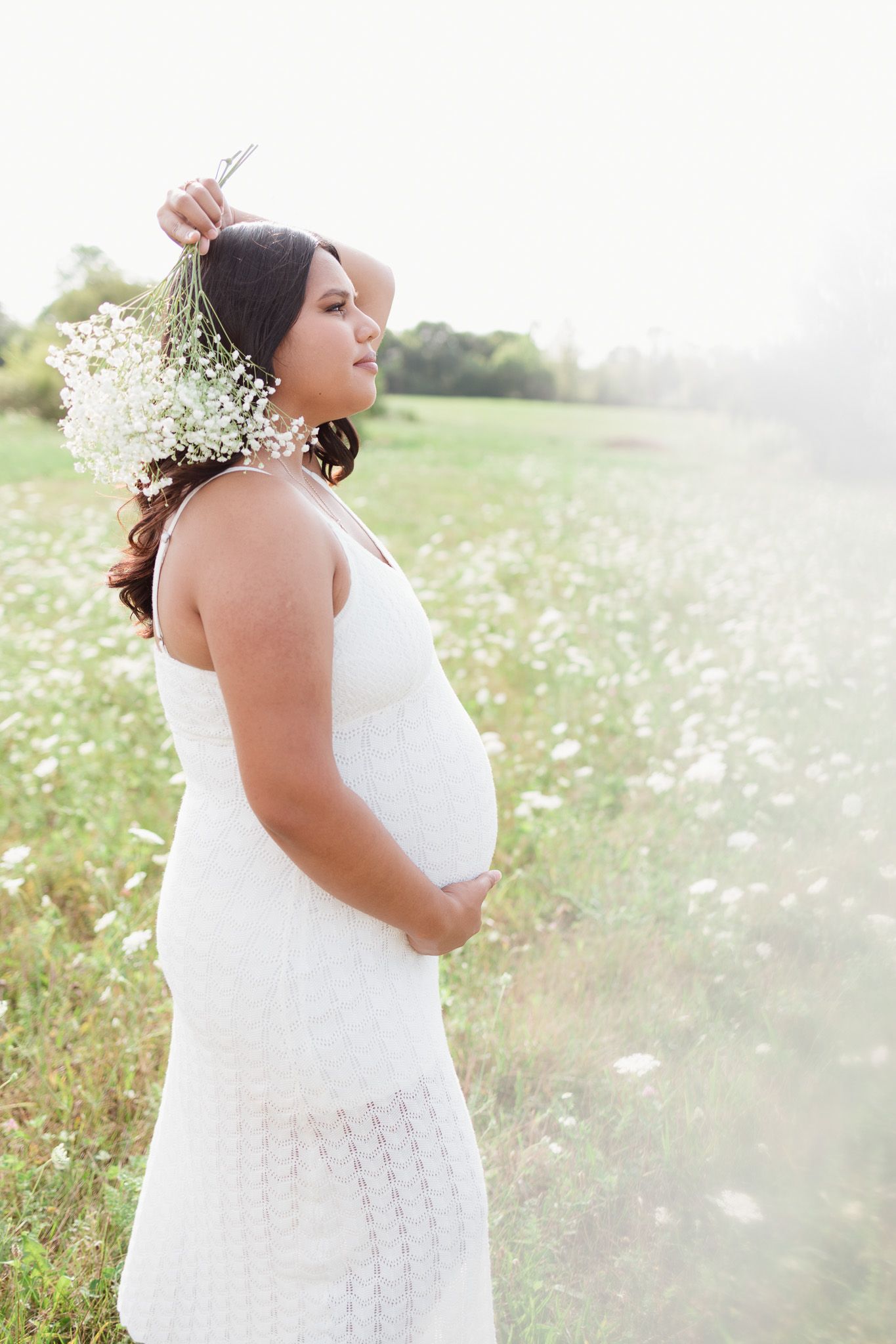 Pregnant woman in white lace dress poses in field of wildflowers at golden hour.