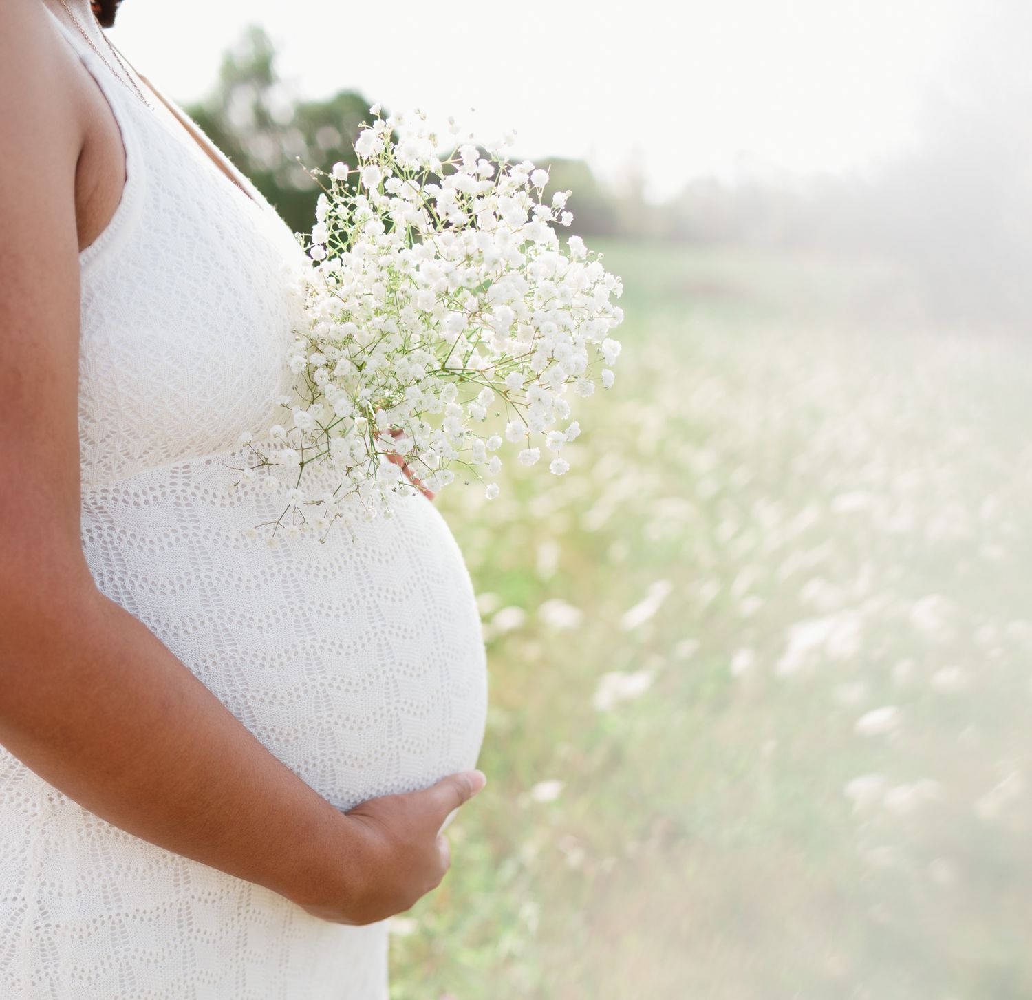 A delicate baby's breath bouquet held against a white maternity dress in a sunlit meadow.