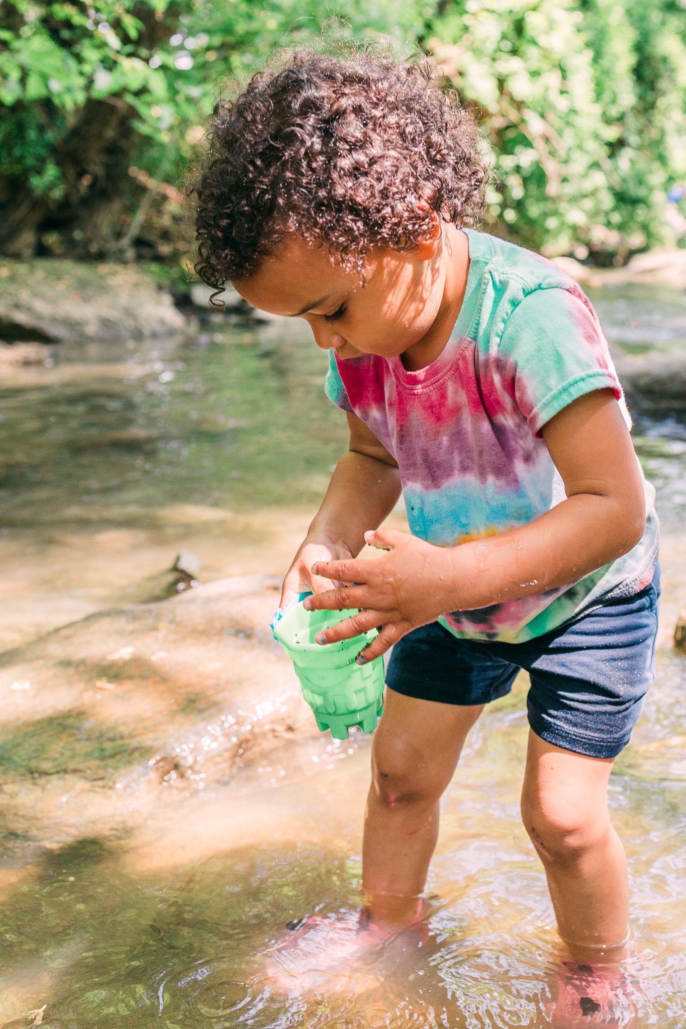 A child wearing a tie-dye shirt and shorts plays in a shallow stream with a green bucket during a sunny day.