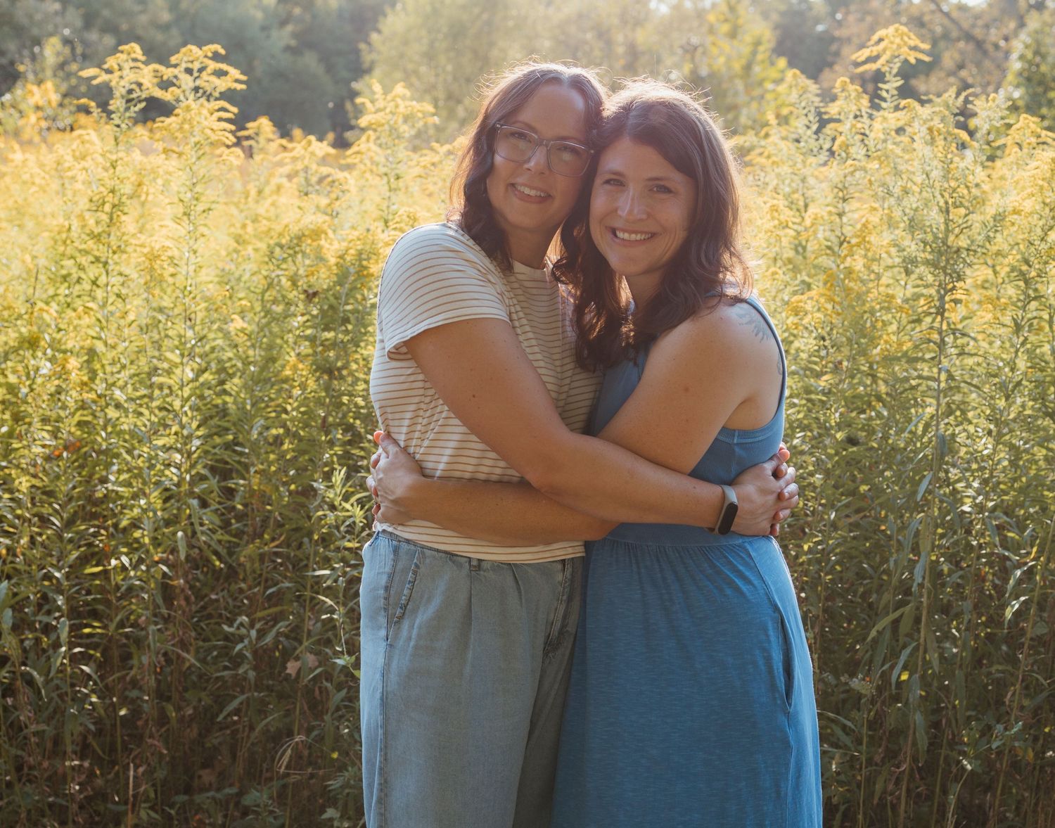 Two people embrace warmly while standing in a field of golden wildflowers at sunset.
