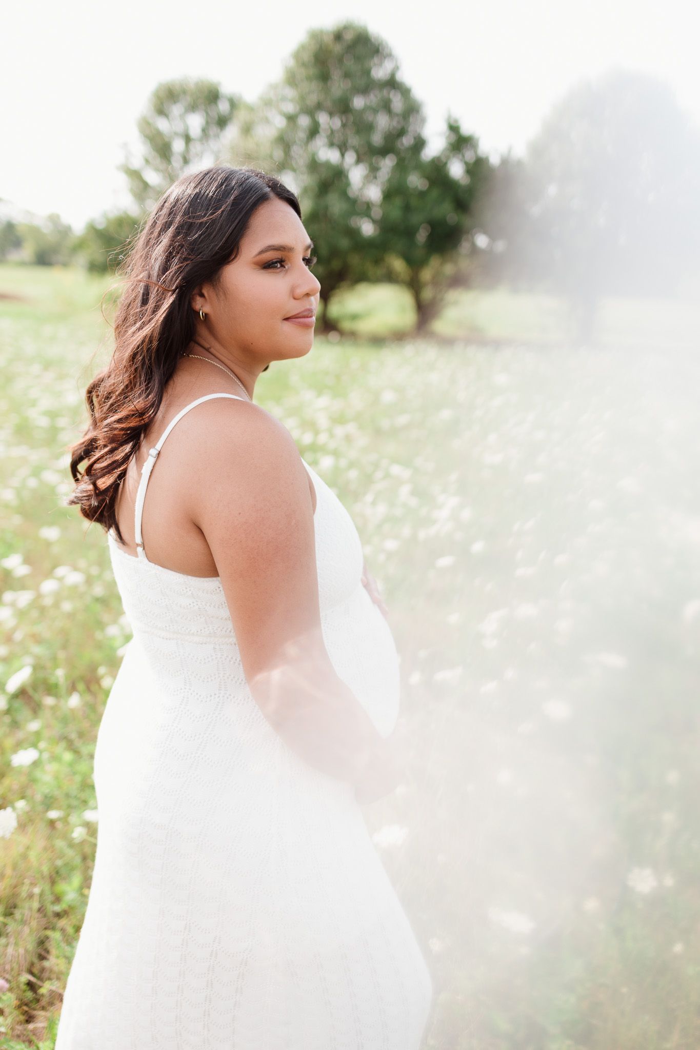 Side profile of pregnant woman in white dress standing in sunny wildflower field.