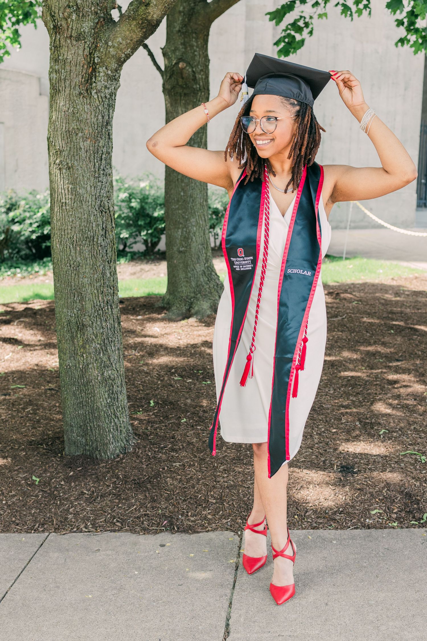 Graduate in cap, gown and red shoes posing playfully near trees.