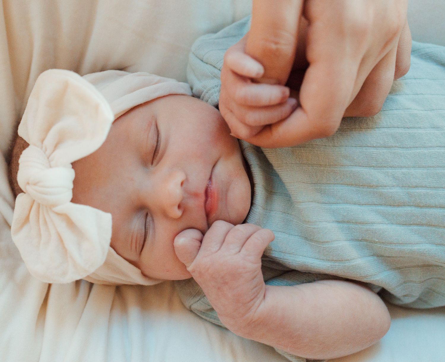 A sleeping infant wearing a cream bow headband rests peacefully on soft fabric while a hand gently touches their face.