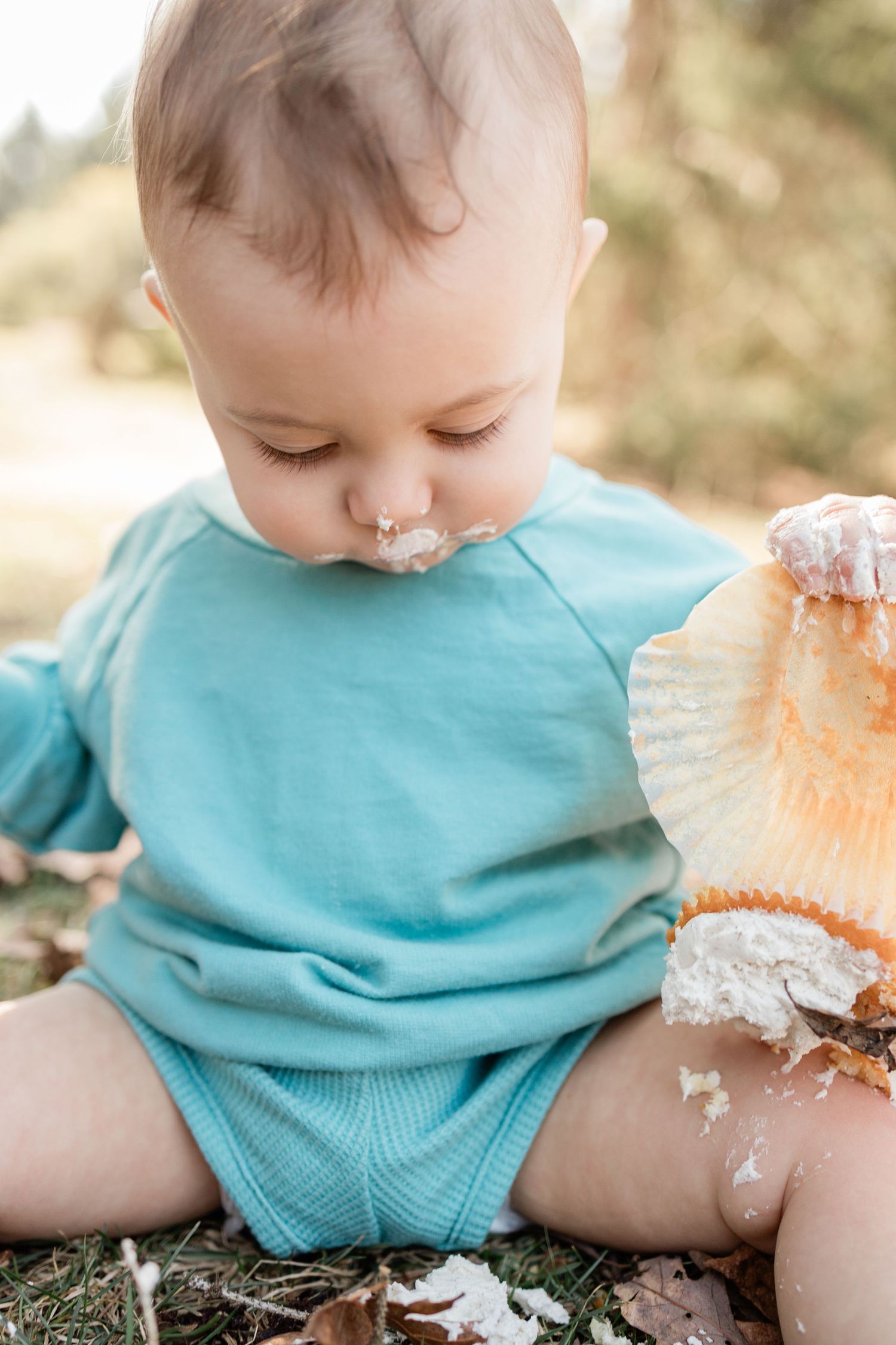 Toddler in teal outfit sitting on ground with crumbs and frosting on face.