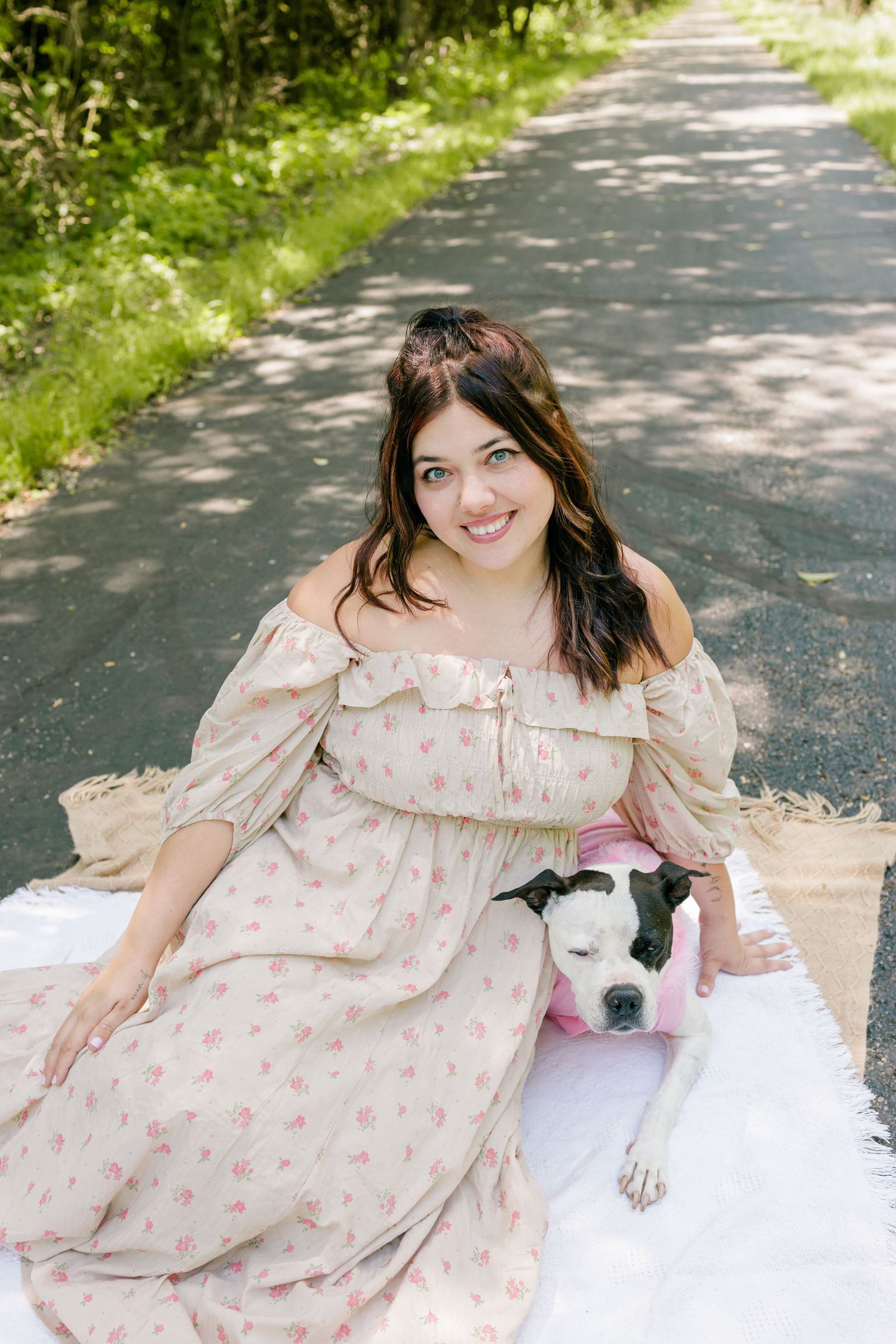 A person in a floral dress sits on a blanket with a black and white dog on a sunny path.