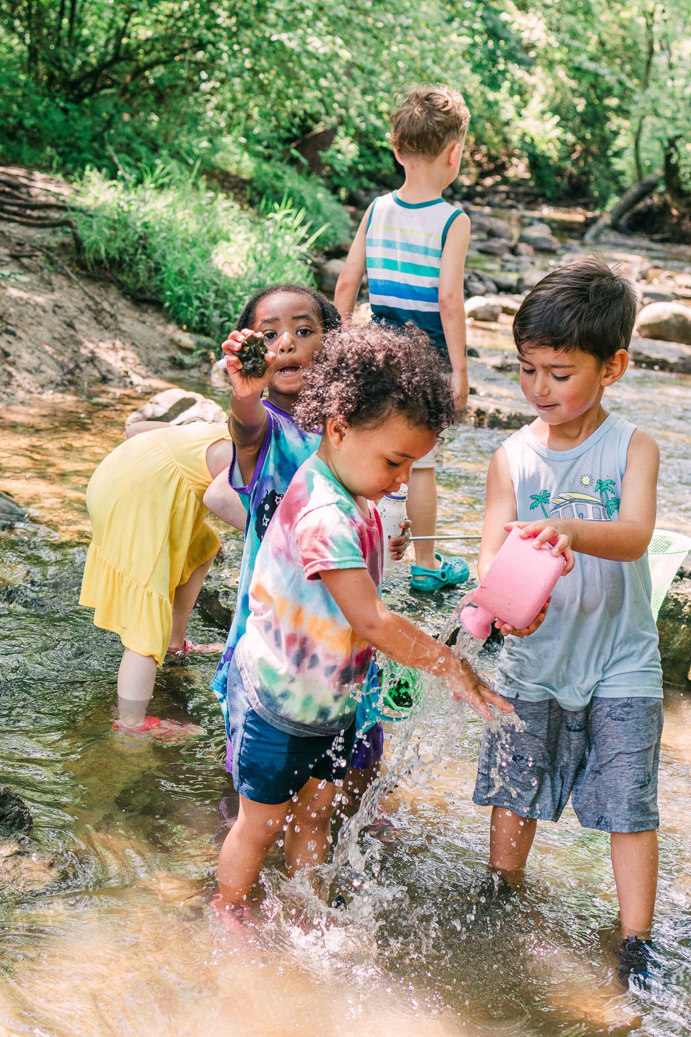 Children playing and splashing in a shallow creek on a sunny summer day surrounded by green trees.