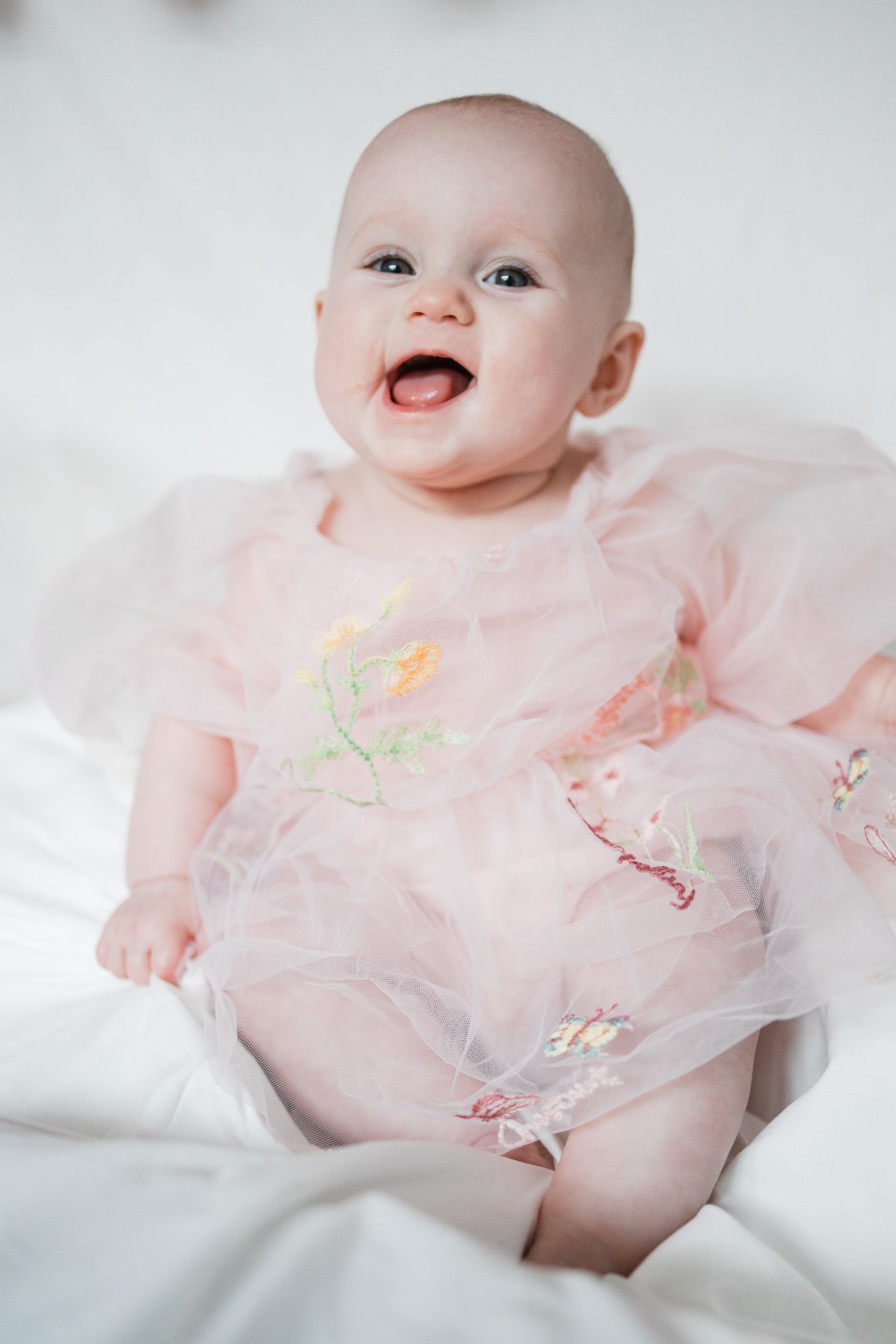 Baby wearing pink tulle dress smiles while laying on white bedding.