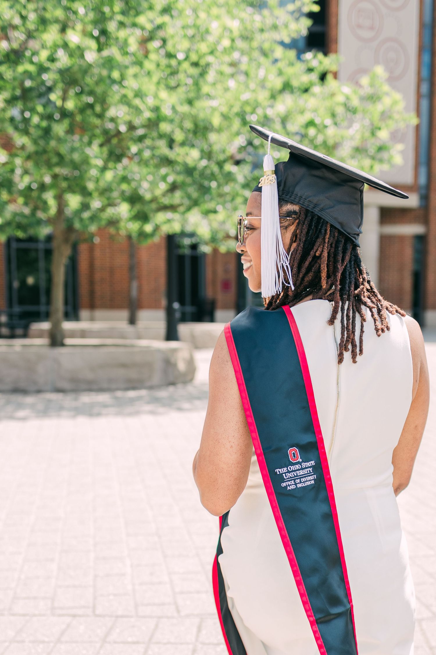 Graduate wearing cap and black and red graduation stole shown from behind on campus.