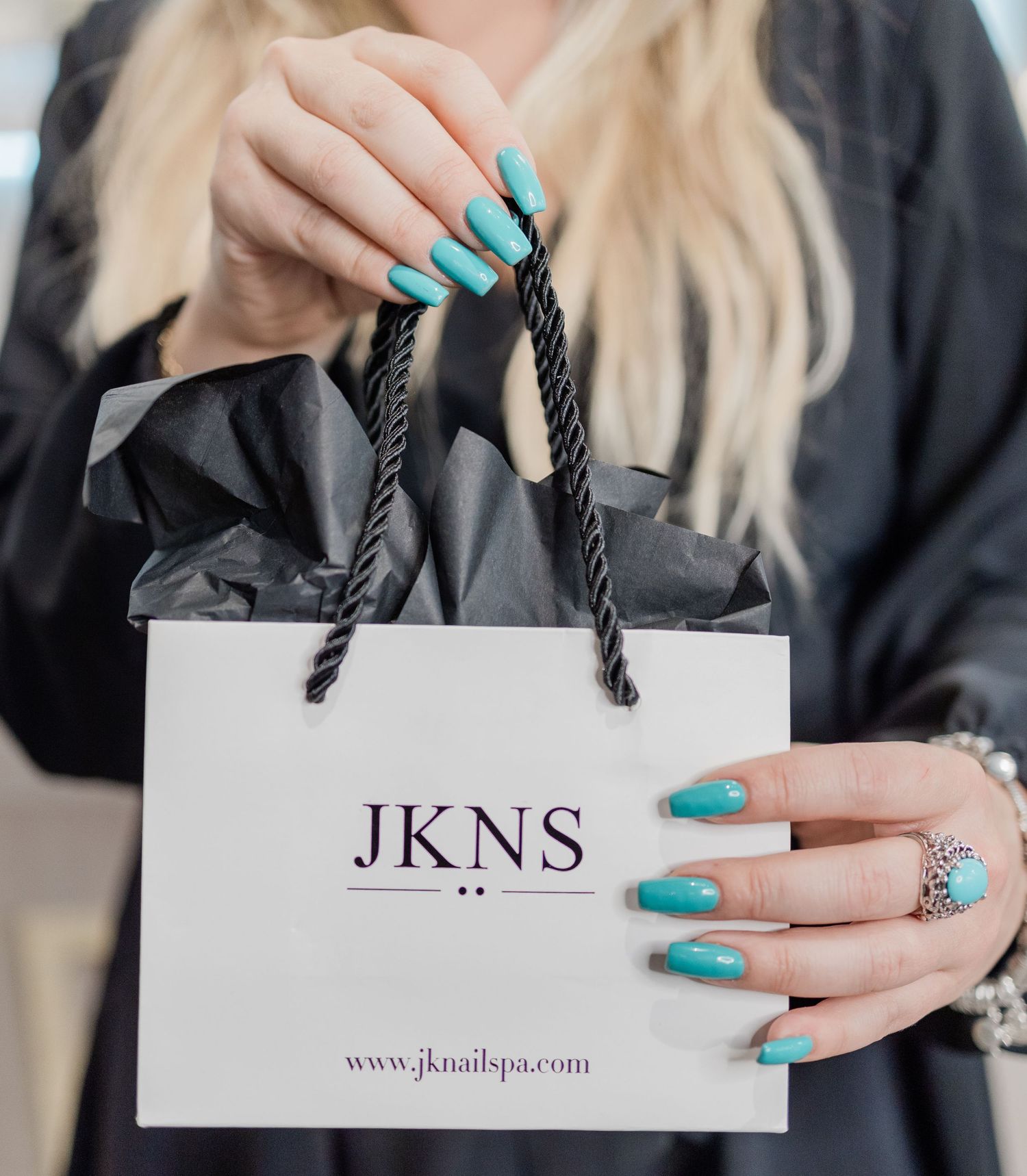 Close-up of turquoise manicured nails holding a white shopping bag with JKNS logo.