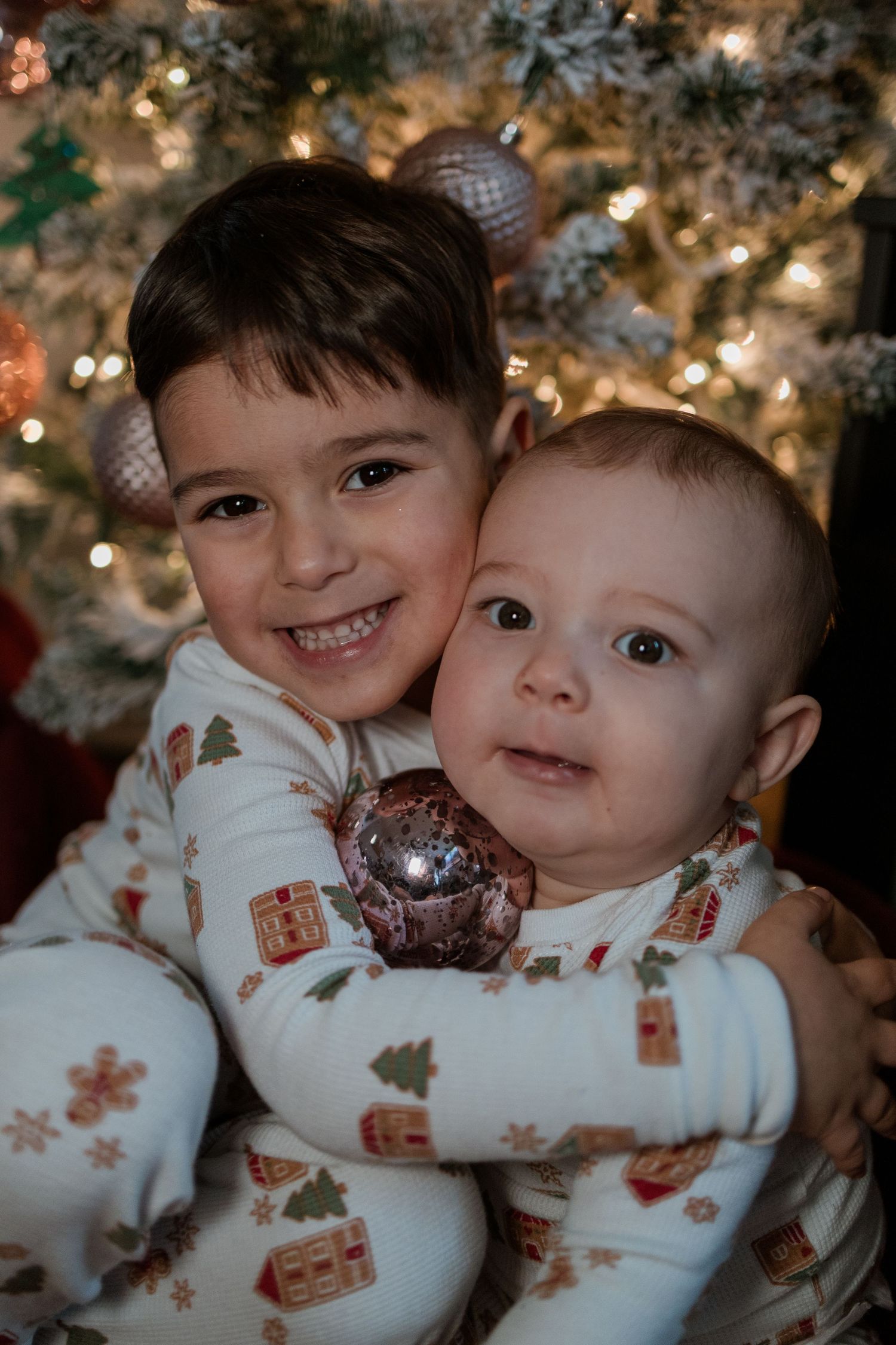 Two children in matching holiday pajamas smile and hug in front of a decorated Christmas tree with twinkling lights.