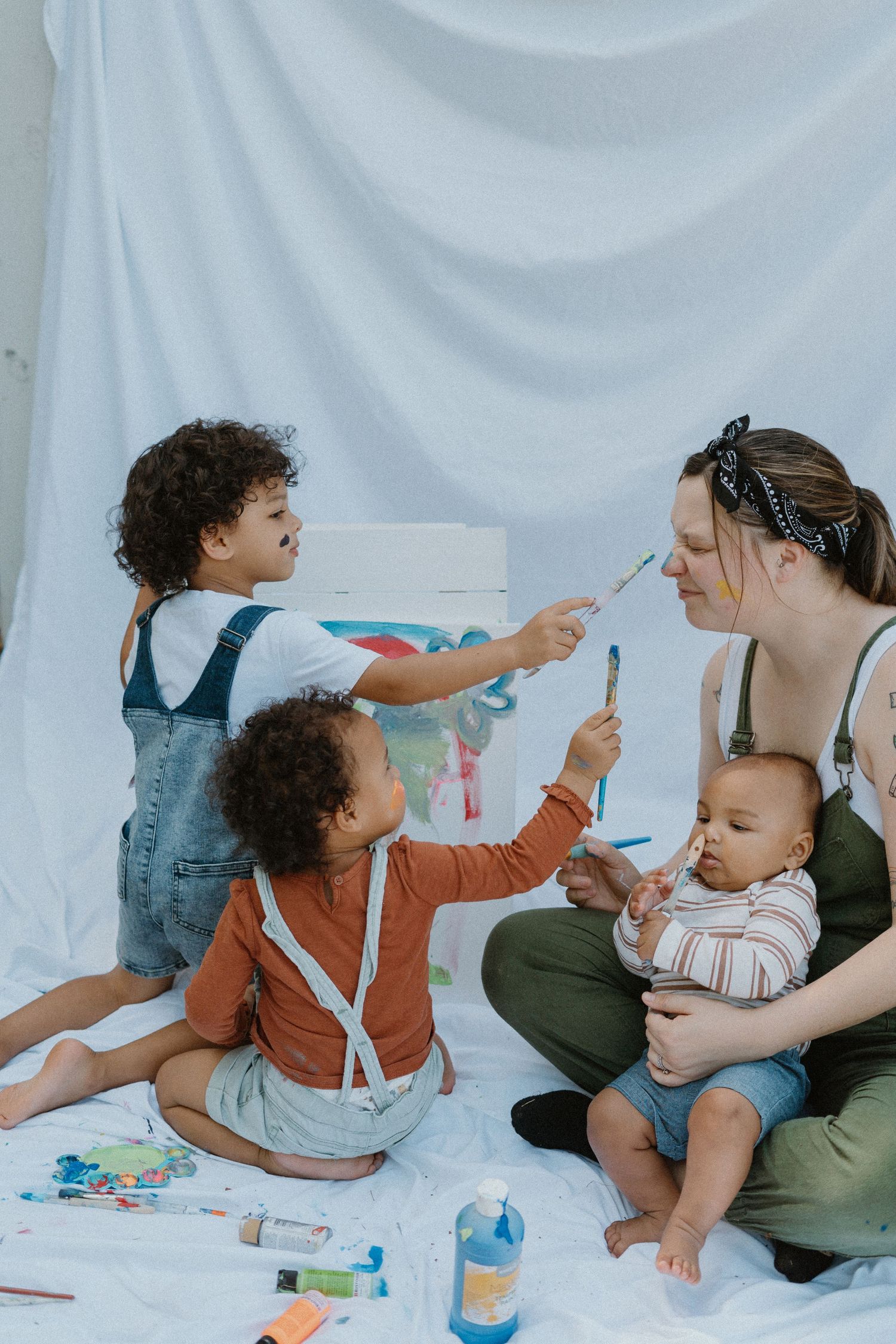 Children painting with brushes while sitting on white backdrop with caregiver and baby.