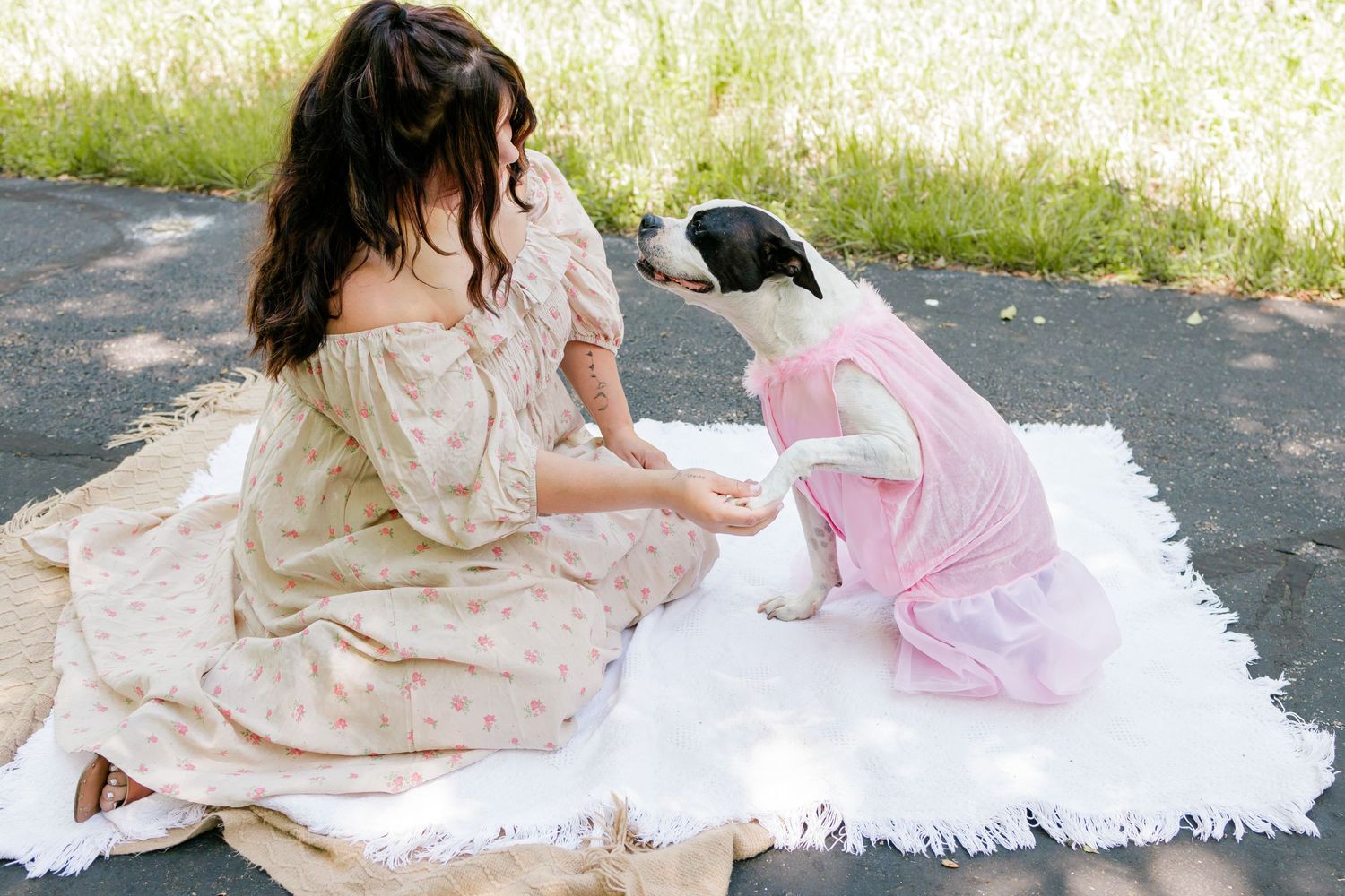Person in floral dress sits on white blanket with black and white dog wearing pink tutu.