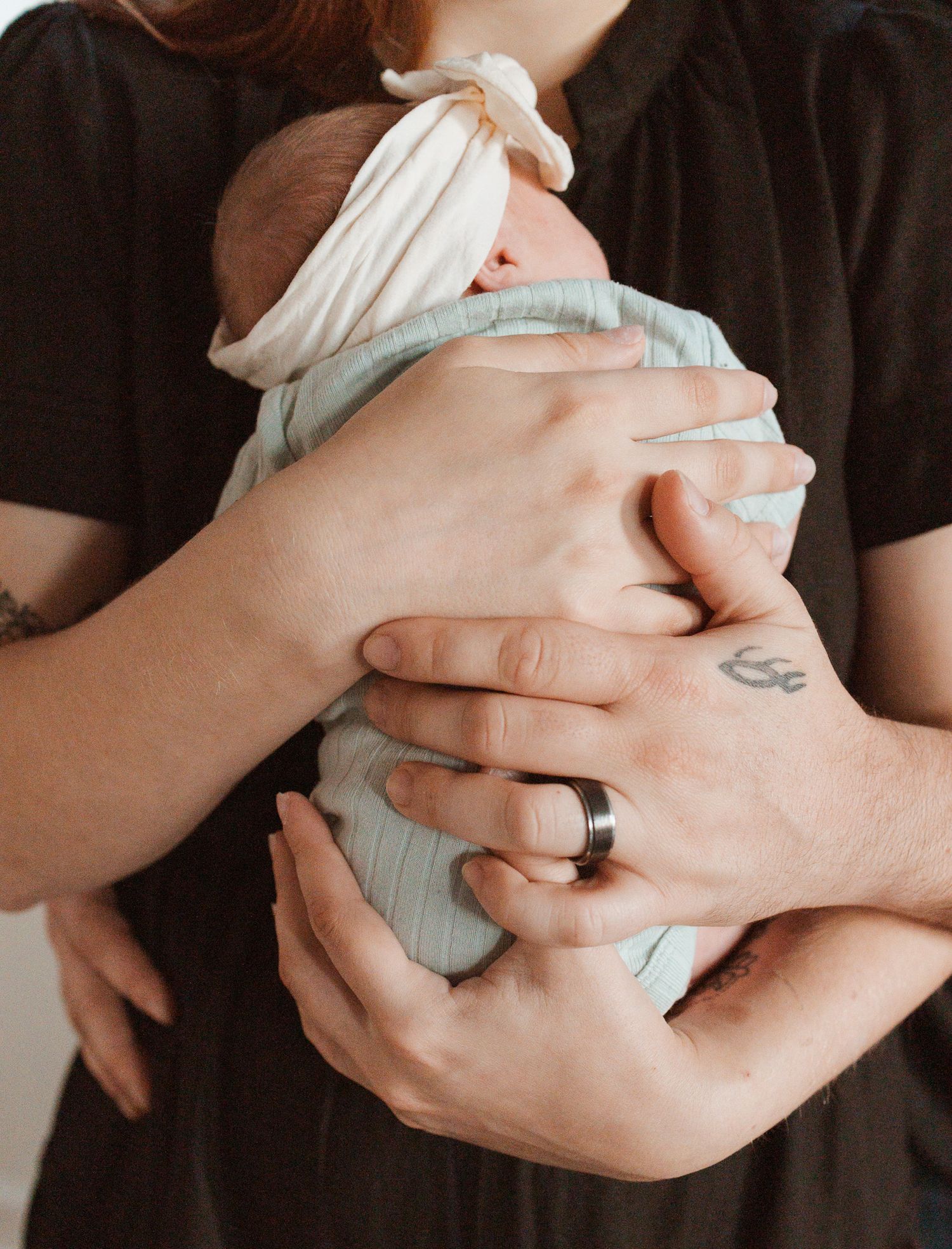 Close-up of hands holding a swaddled newborn baby wearing a mint green outfit, with a visible ring and small tattoo.