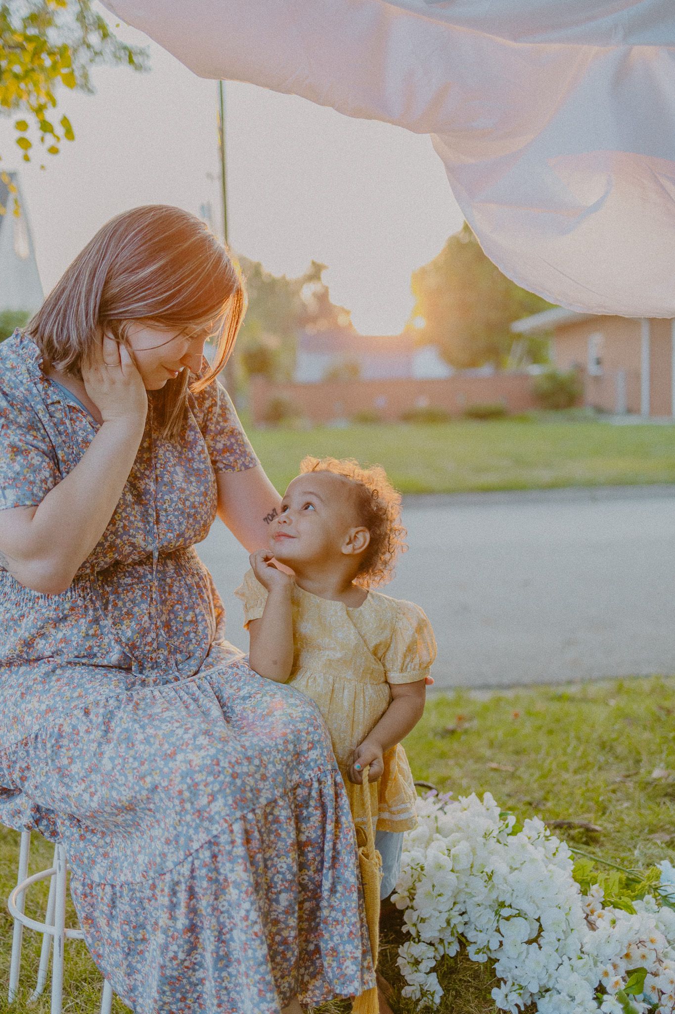 A touching moment between parent and child sitting outdoors at sunset with white flowers nearby.
