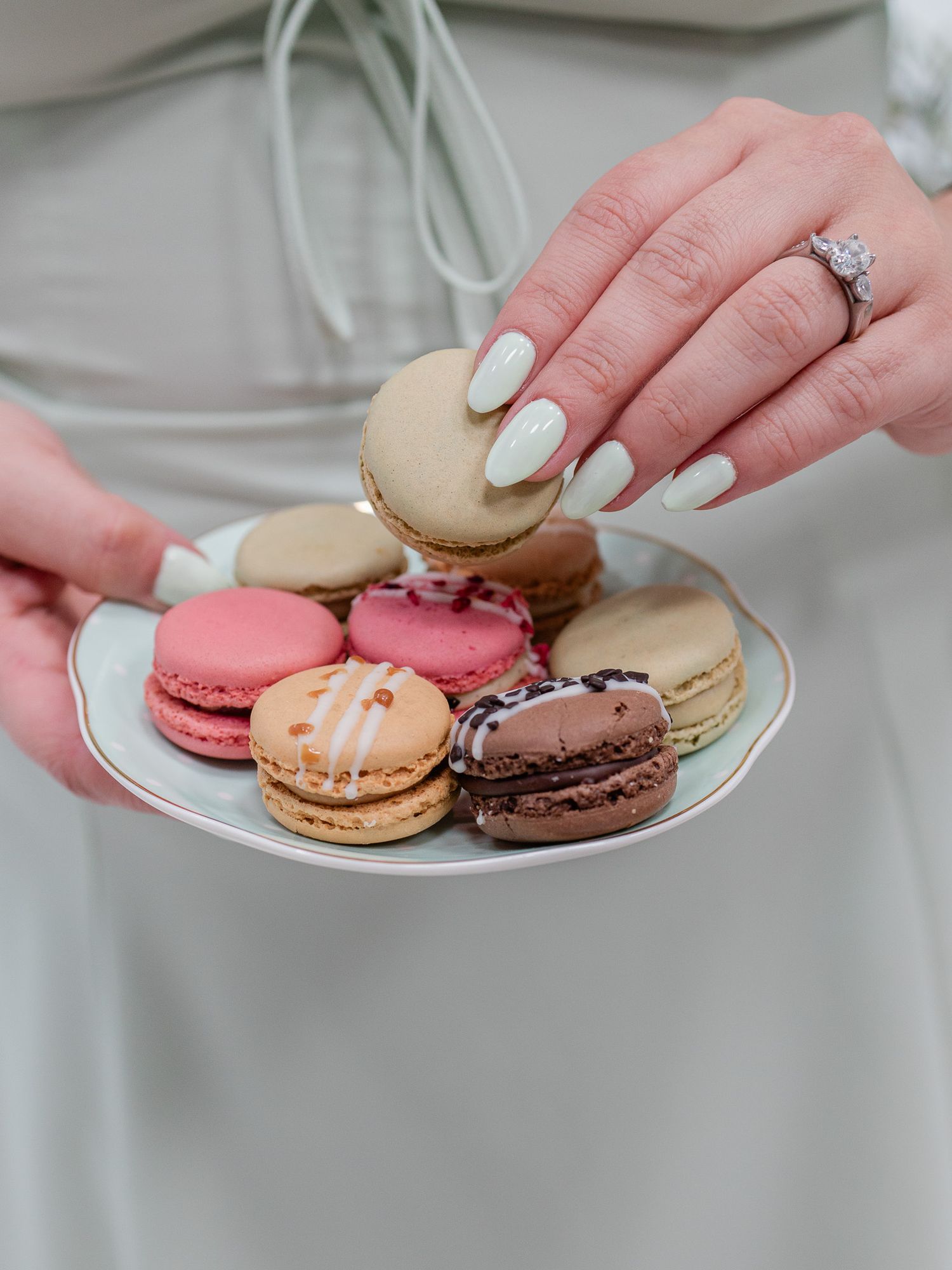 Manicured hands with white nail polish holding colorful French macarons on a plate.