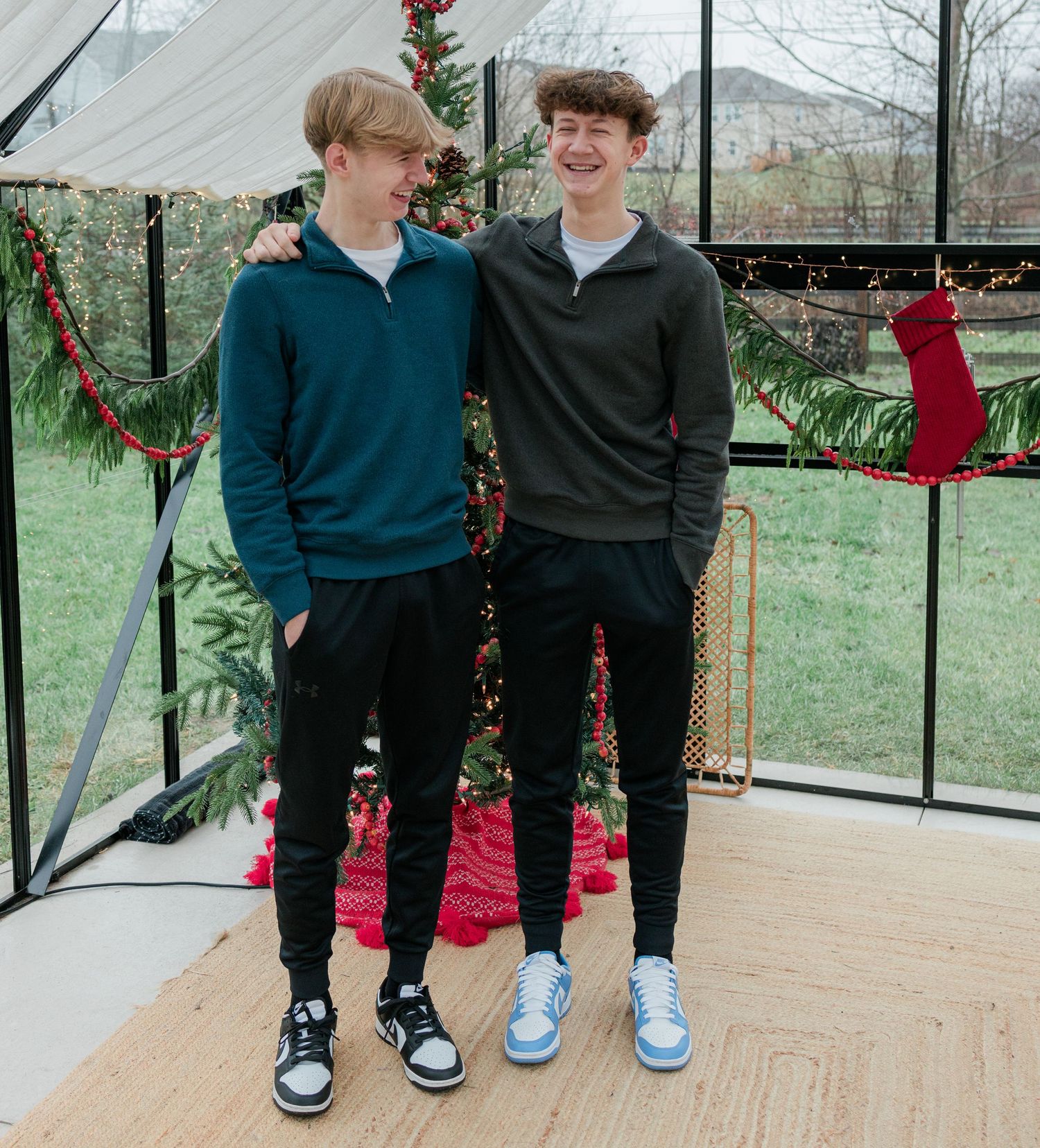Two young men in casual sweaters stand together by a Christmas tree with holiday decorations.