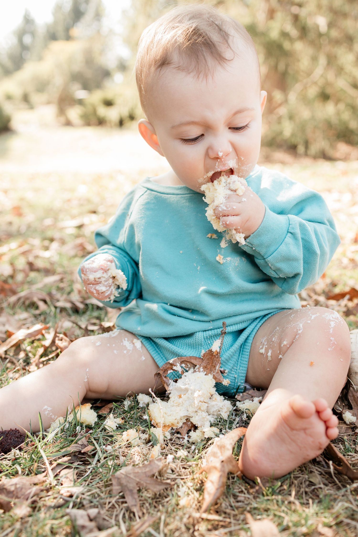Baby in teal outfit sitting on ground messily eating cake.
