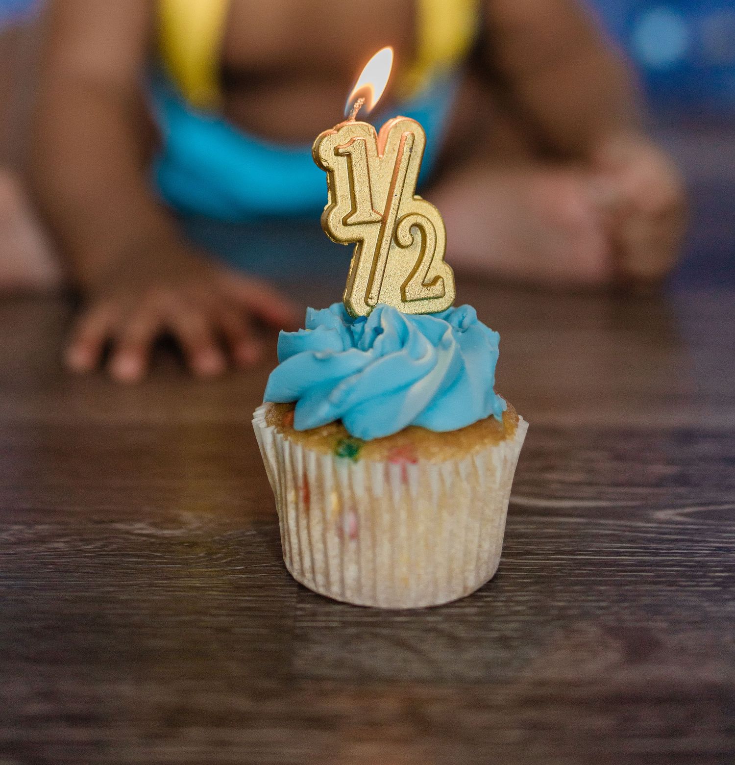 Cupcake with blue frosting and lit half birthday candle on wooden table.