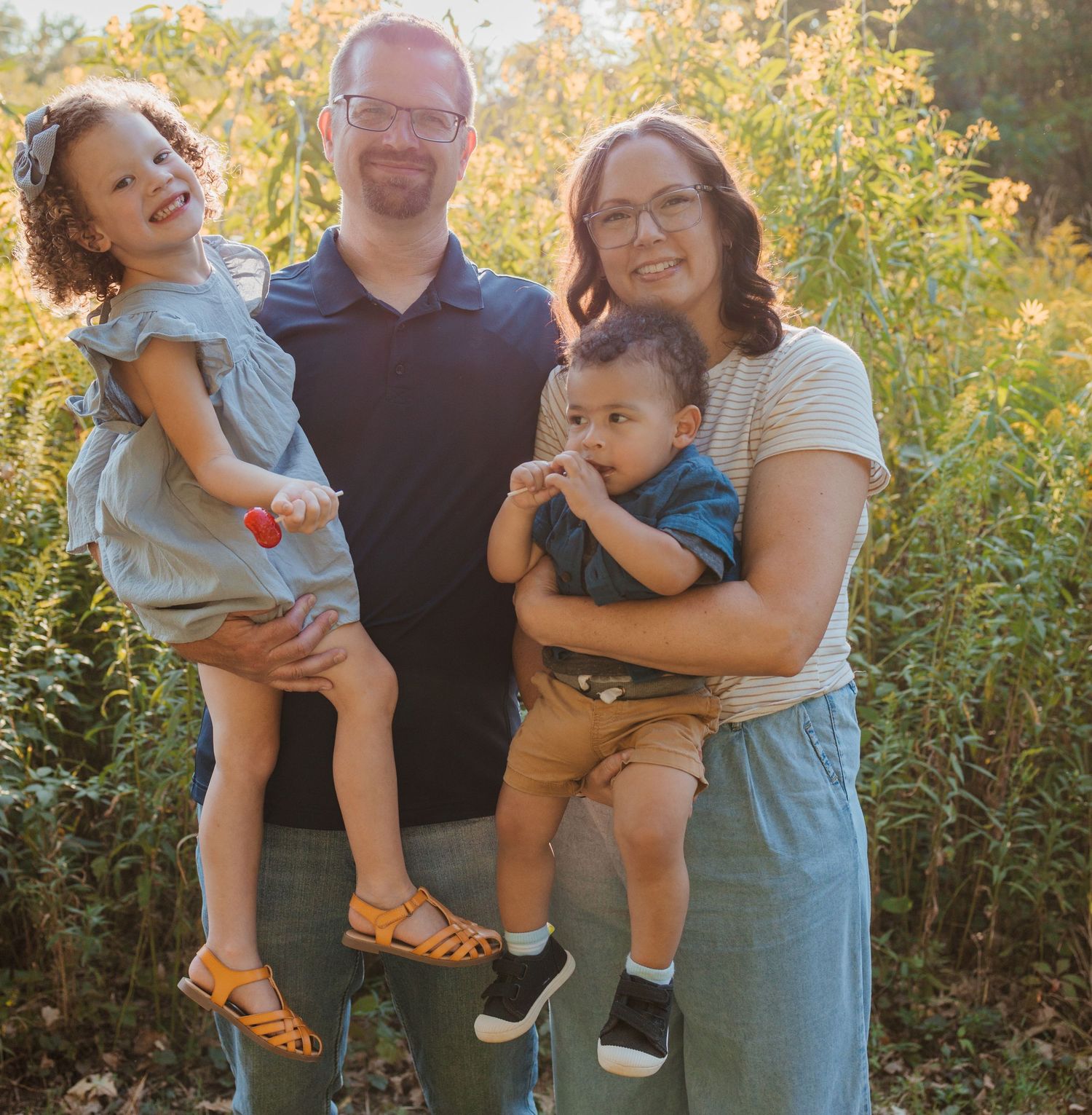 A joyful family portrait outdoors with two adults and two children surrounded by autumn foliage.
