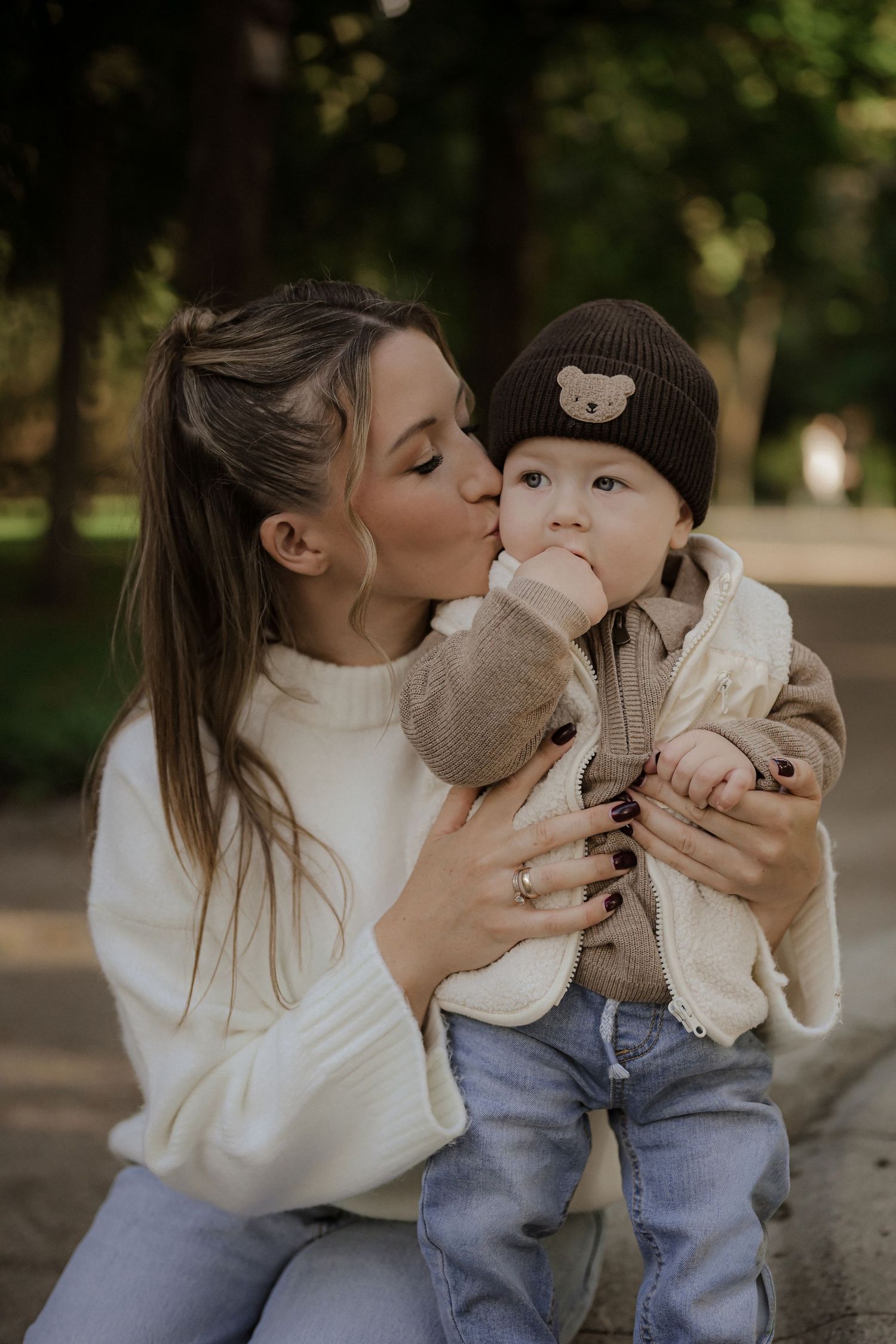 A young mother in a white sweater tenderly kisses her baby wearing a brown bear hat in a park setting.