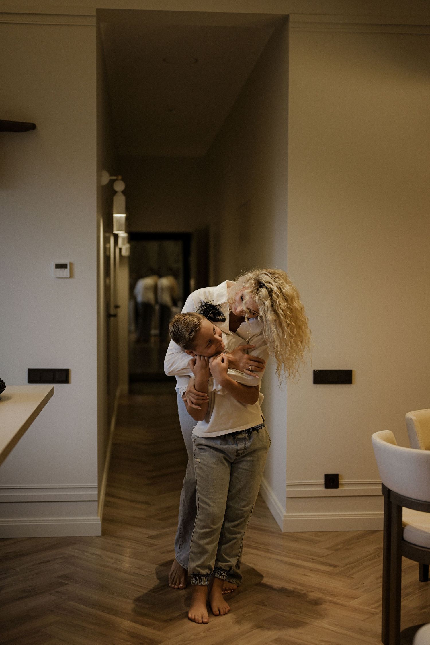 A woman with curly blonde hair hugs and kisses a child in a modern home hallway with herringbone wood floors.